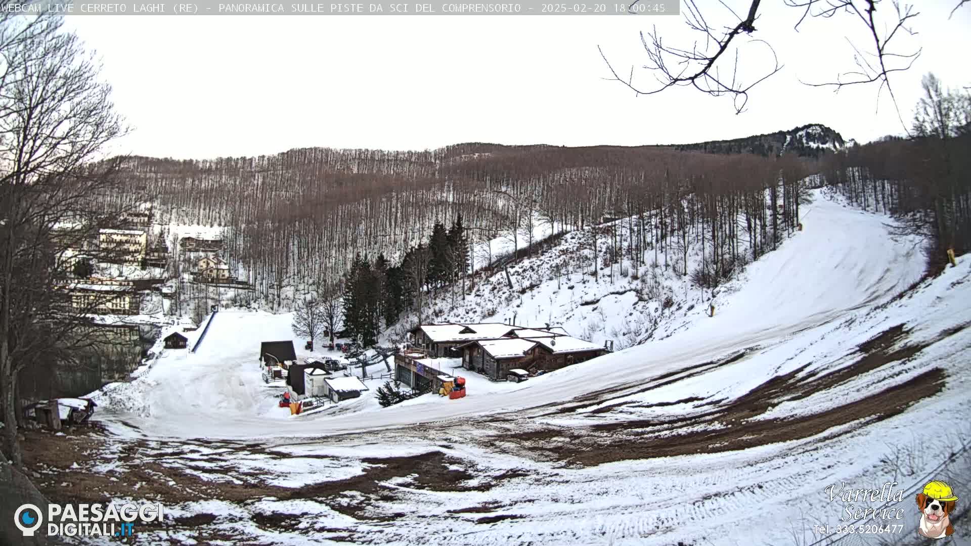 A snowy ski slope with several buildings at its base, nestled among a hillside of leafless trees under a mostly clear sky.
