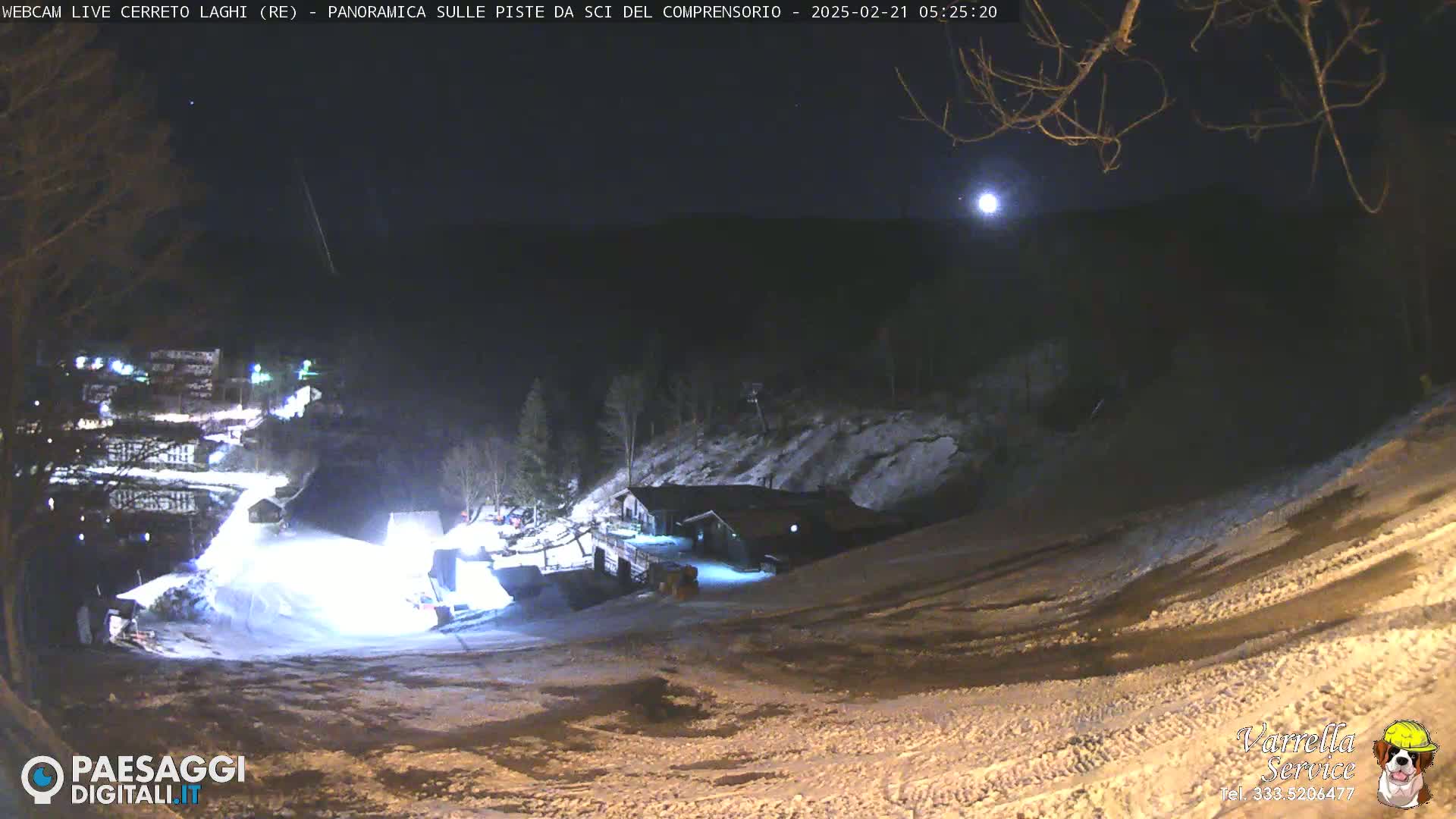 A nighttime view of a snow-covered ski slope illuminated by artificial lights under a mostly clear sky with a bright moon.