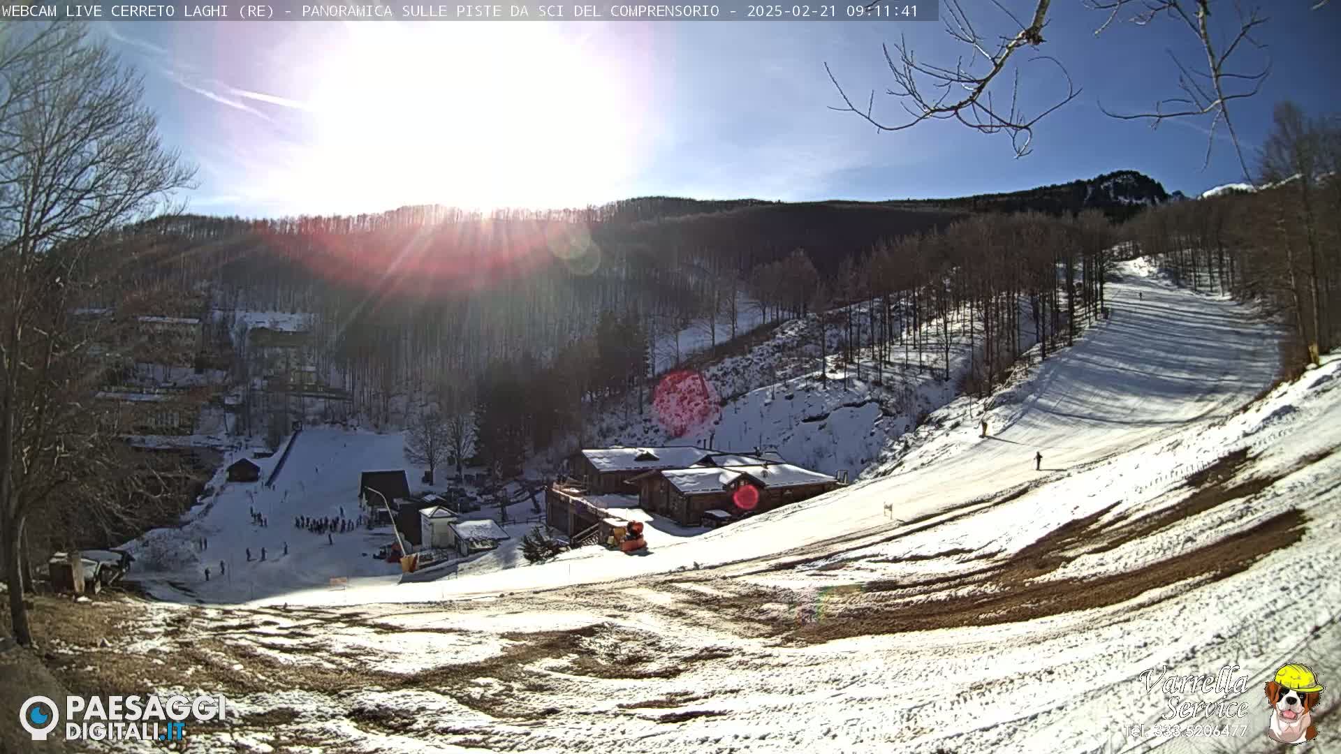 A sunny, snow-covered ski slope with a few skiers and a building at its base, nestled among bare trees on a hillside.