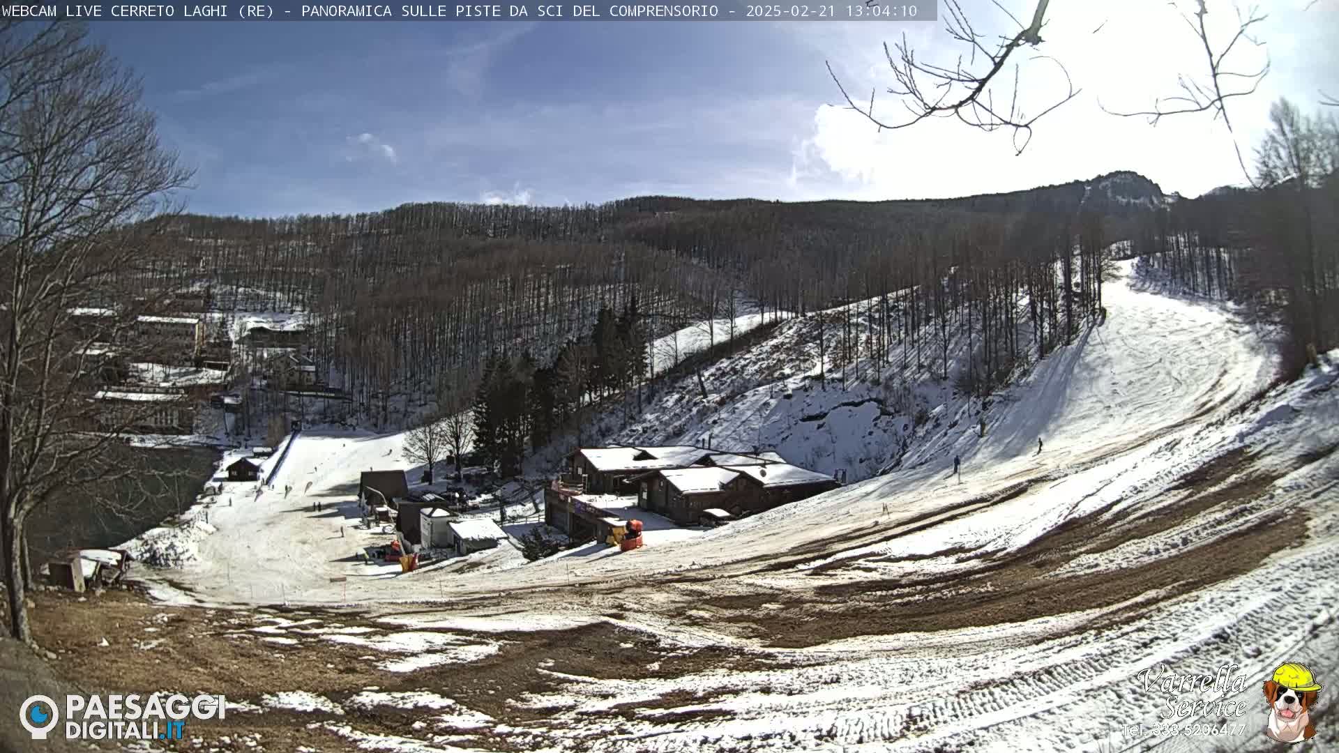 A snow-covered ski slope with several buildings at its base, under a mostly sunny sky.