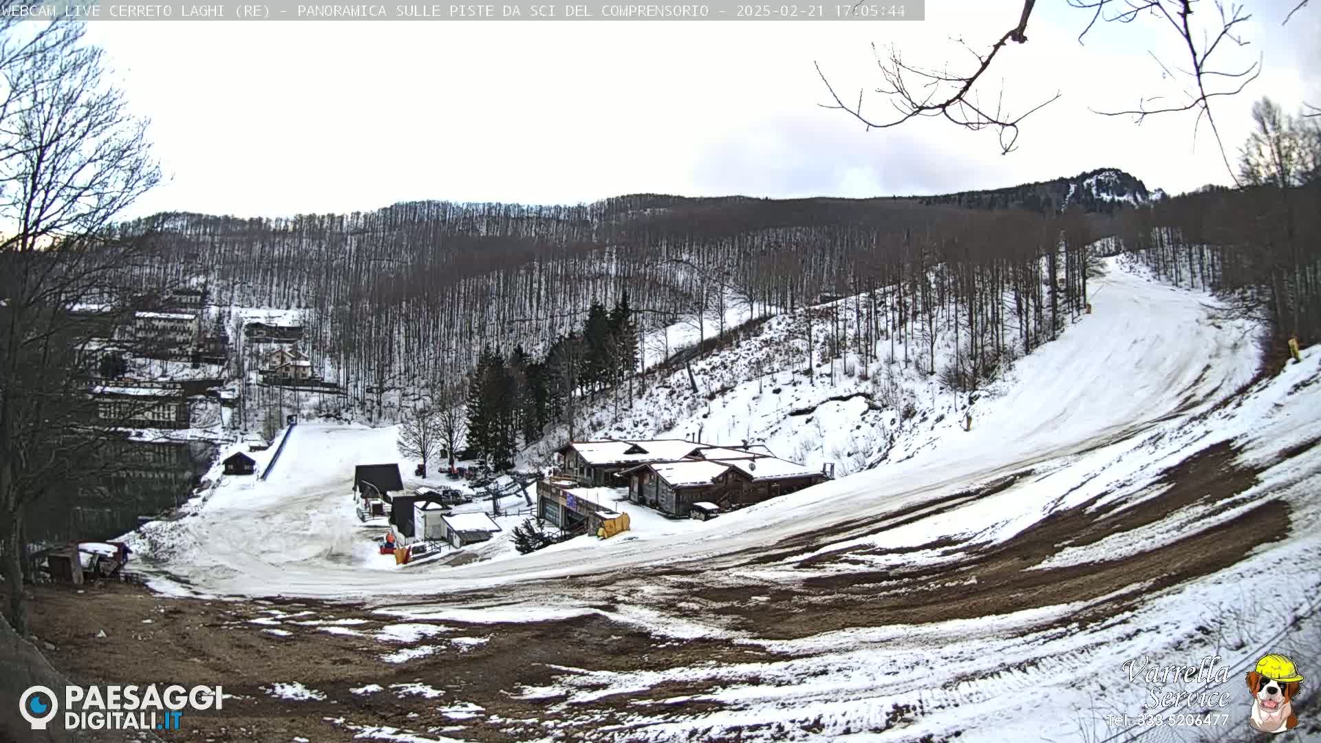 A snow-covered ski slope with several buildings and a forest in the background under a partly cloudy sky.