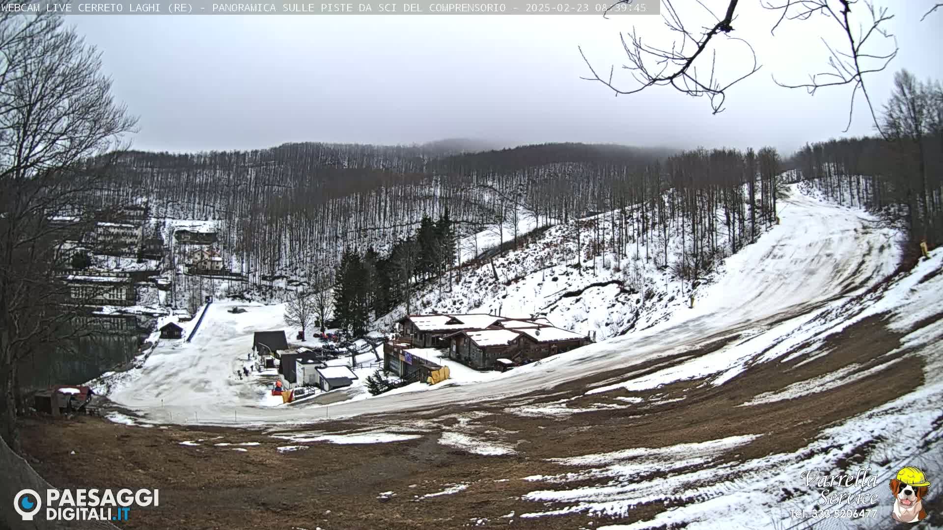 A snow-covered ski slope descends from a cluster of buildings nestled among bare, snow-dusted trees under an overcast sky.