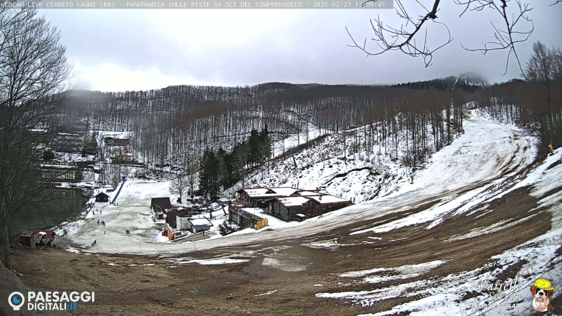A mostly cloudy day reveals a snow-covered ski slope with a few buildings at its base, surrounded by a treeline.