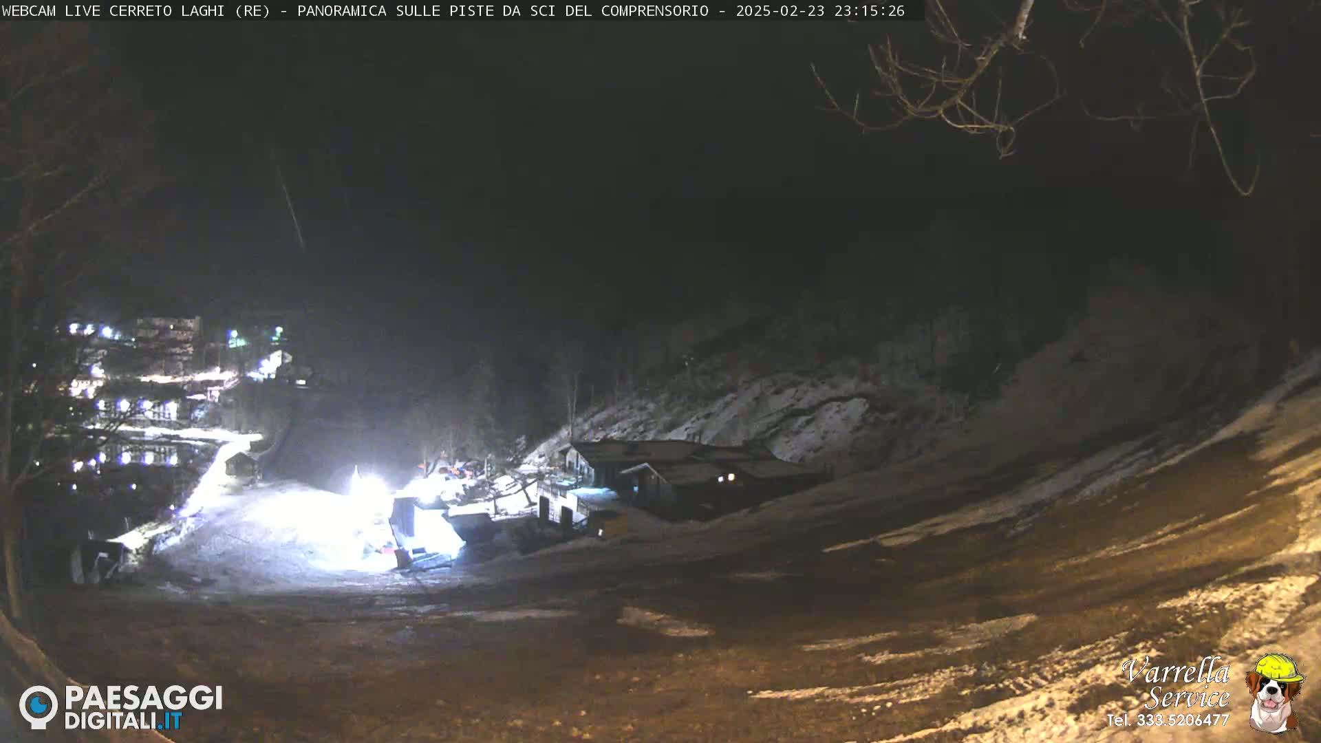 A nighttime, snow-covered ski slope is illuminated by lights, showing a building and some activity near the bottom of the hill.