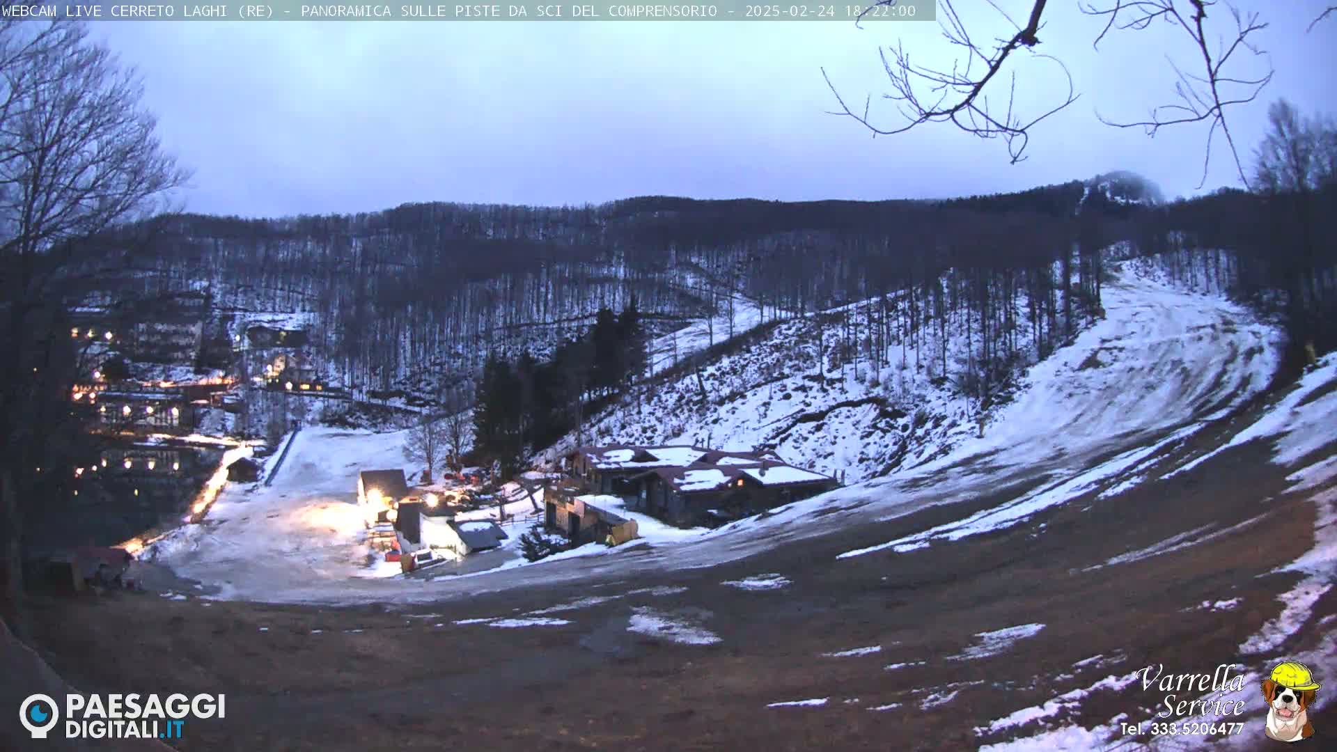 A nighttime view of a snow-covered ski slope with a few buildings illuminated at the bottom of the hill under a twilight sky.