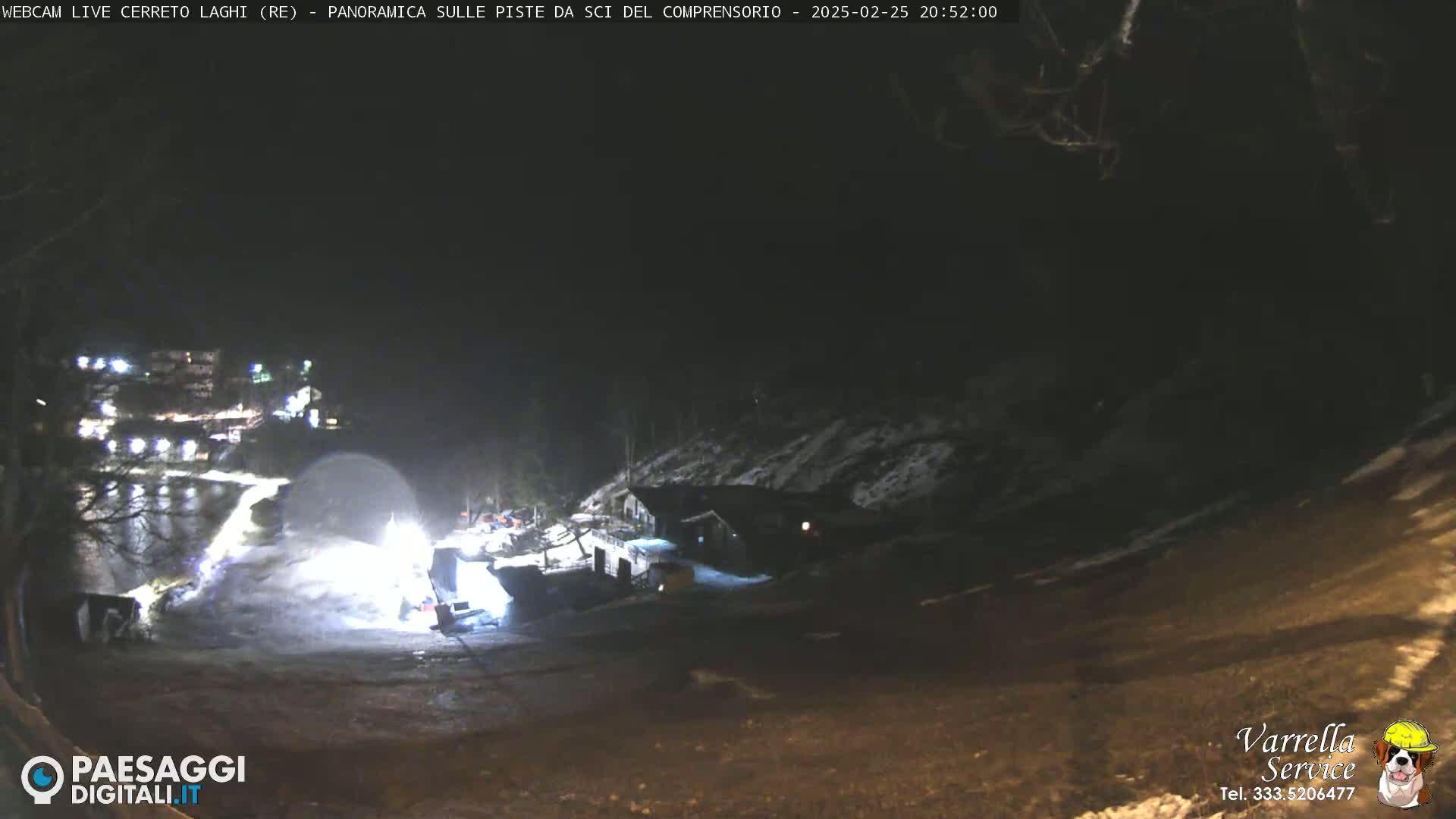 A nighttime, snow-covered landscape is illuminated by artificial lights, showing a building and some vehicles near a sloped area.