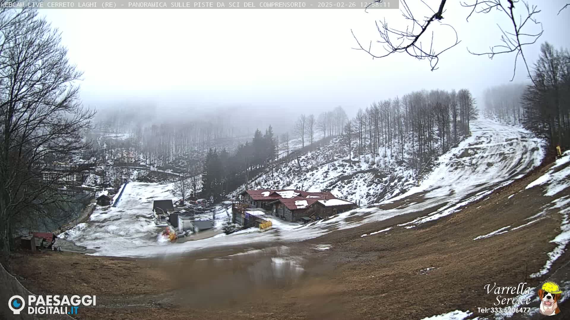 A foggy, snowy hillside scene shows a ski resort with buildings, a mostly snow-covered ski slope, and bare trees.