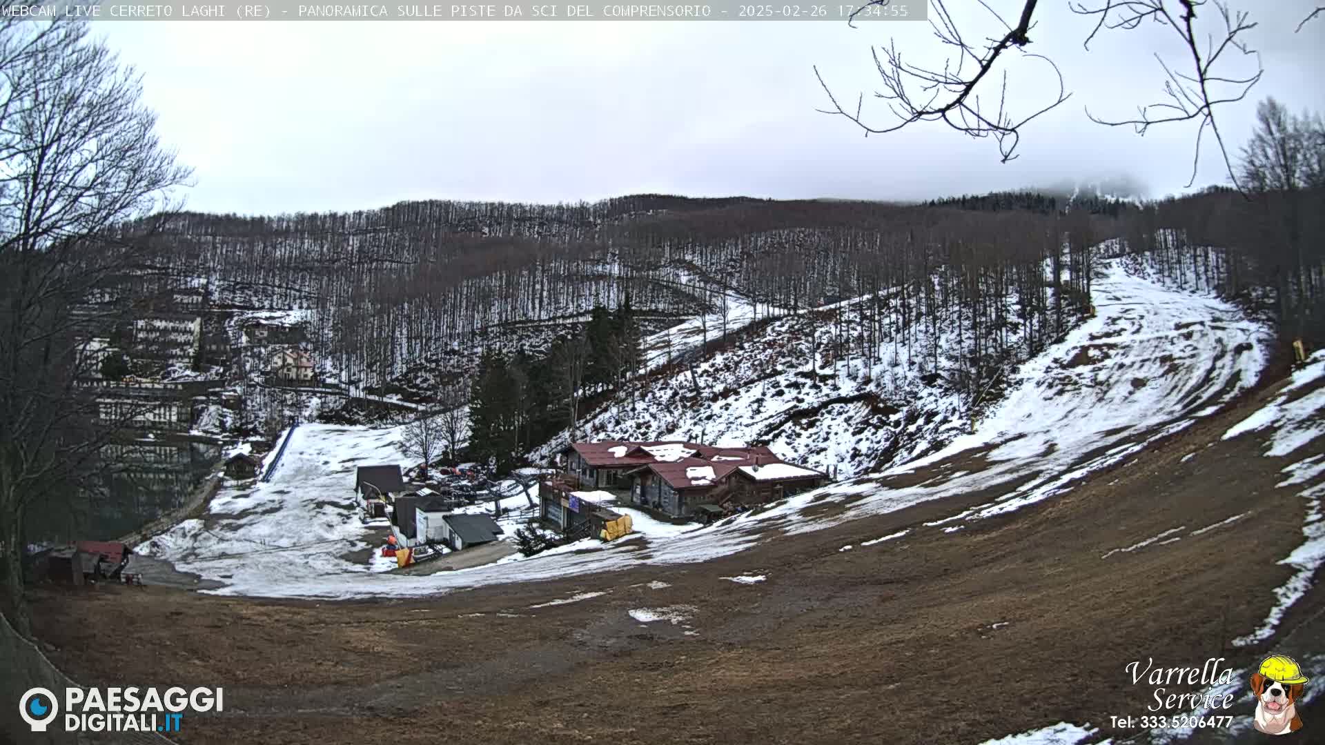 A snow-dusted ski slope descends toward a cluster of buildings nestled amongst bare, snow-covered trees under an overcast sky.