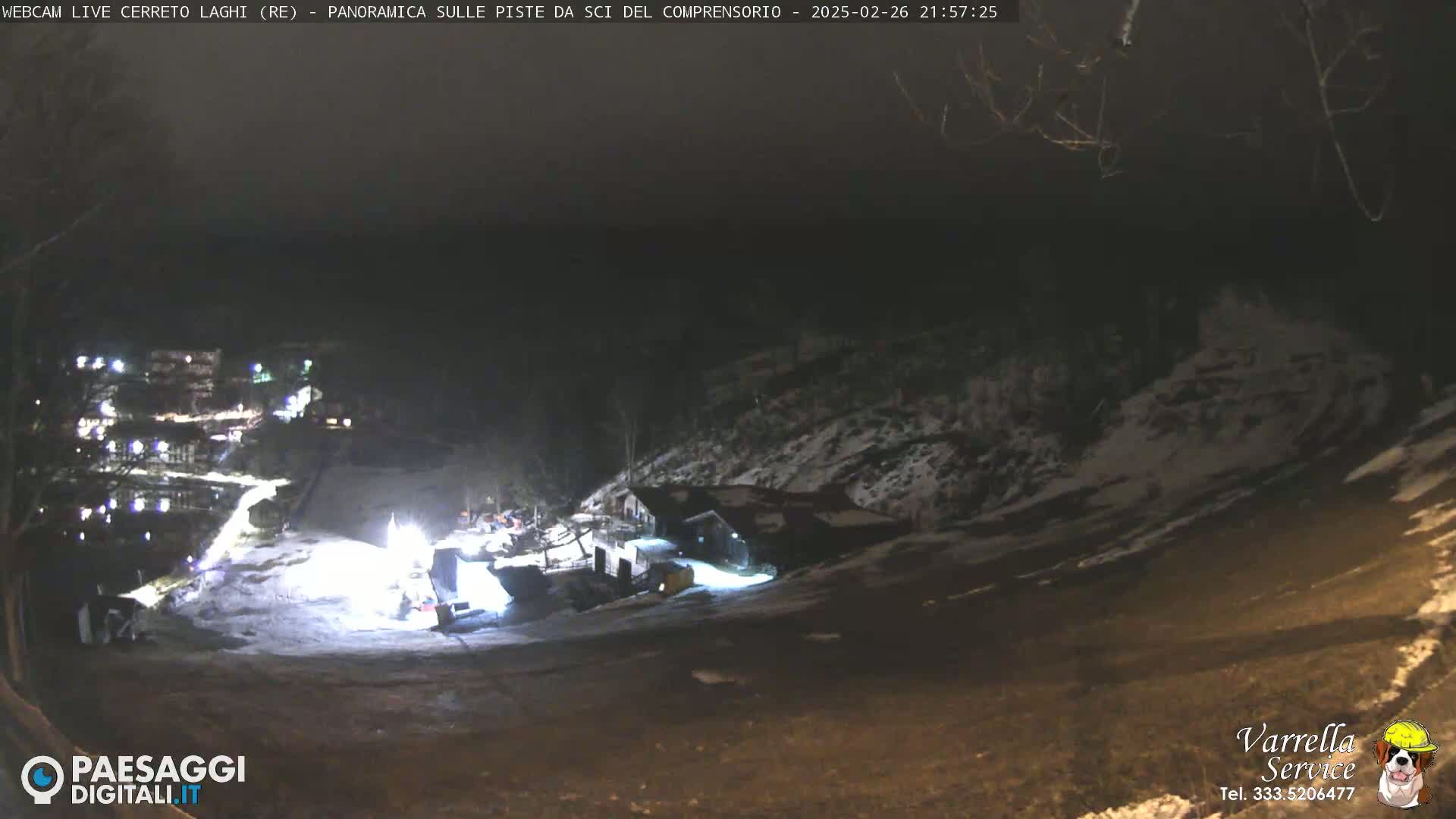 A nighttime, snow-dusted hillside scene shows illuminated buildings and a snow-covered slope with some vehicles near the bottom.