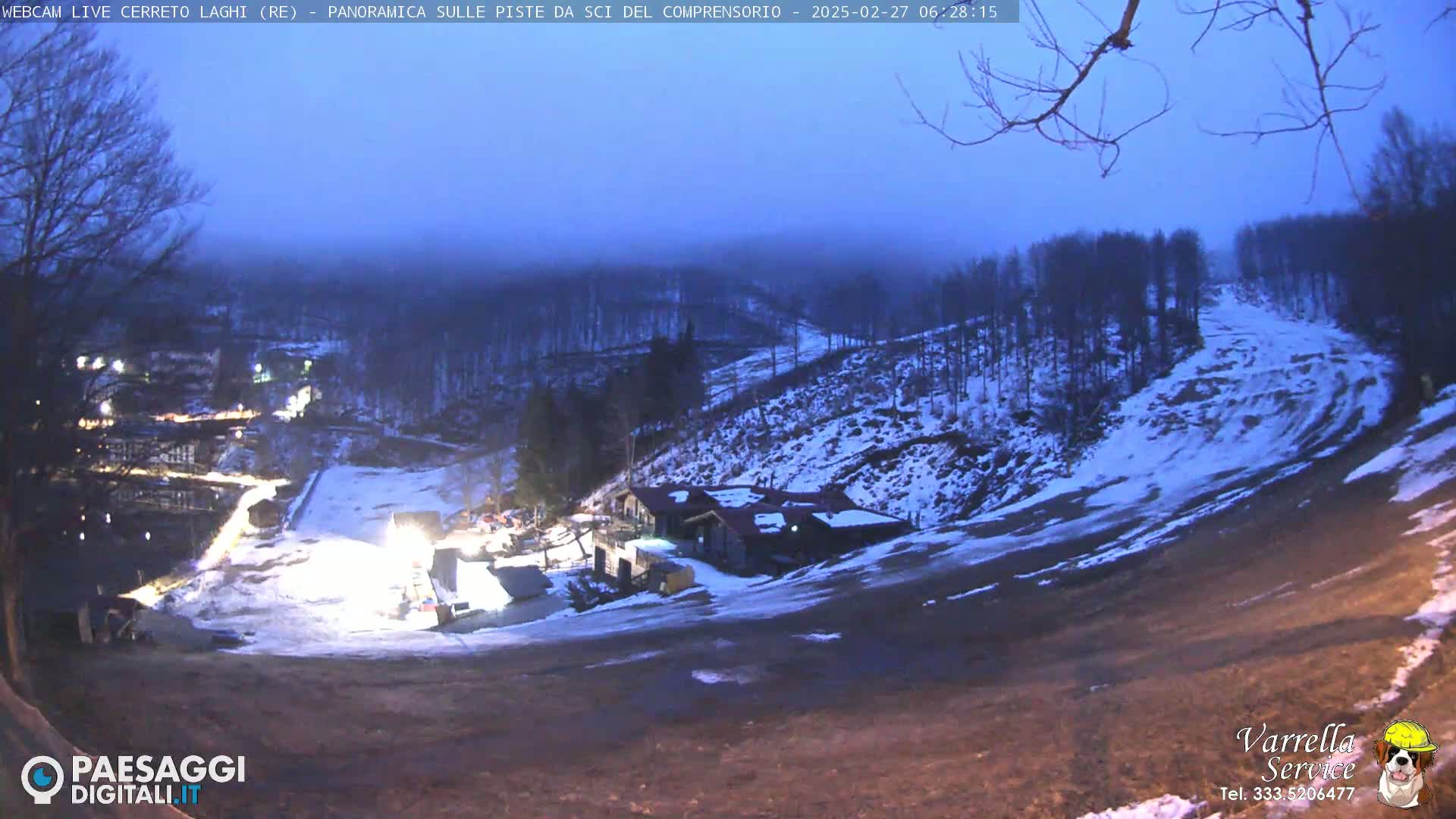 A snow-covered ski slope at twilight, with a building and lights visible in the lower middle ground, under a hazy, dark blue sky.
