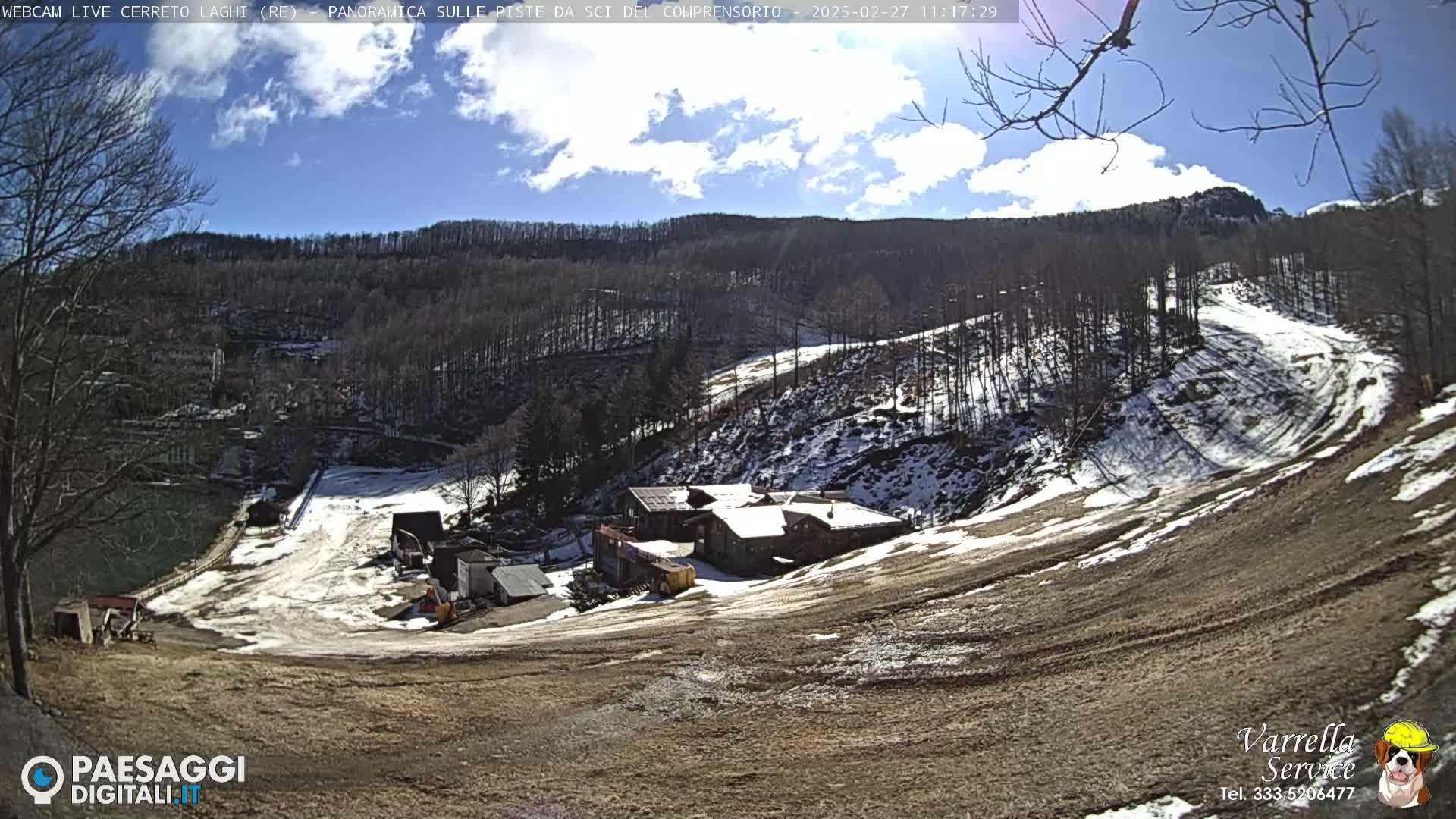 A mostly sunny day reveals a snow-dusted ski slope with a cluster of buildings near the bottom of the hill, surrounded by leafless trees and a partly cloudy sky.