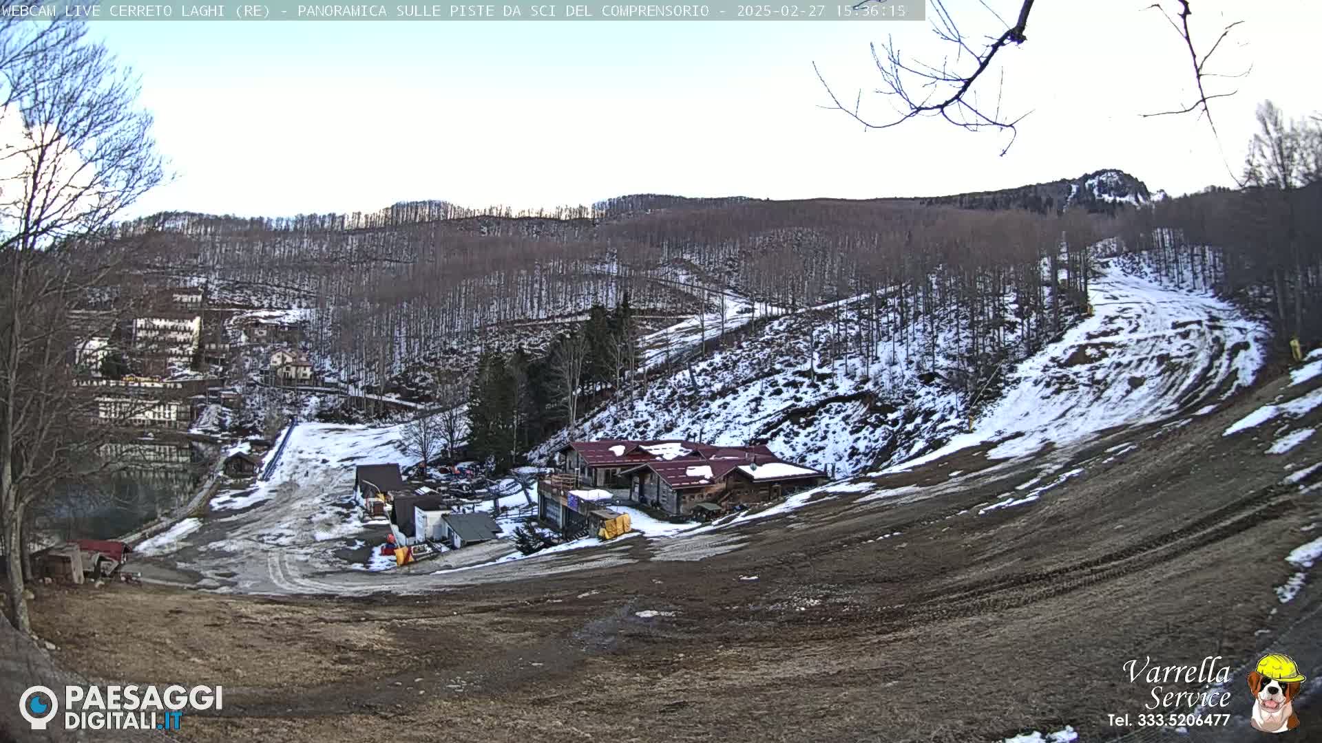 A mostly snow-covered ski slope with a building and some smaller structures at its base, under a mostly clear, bright sky.