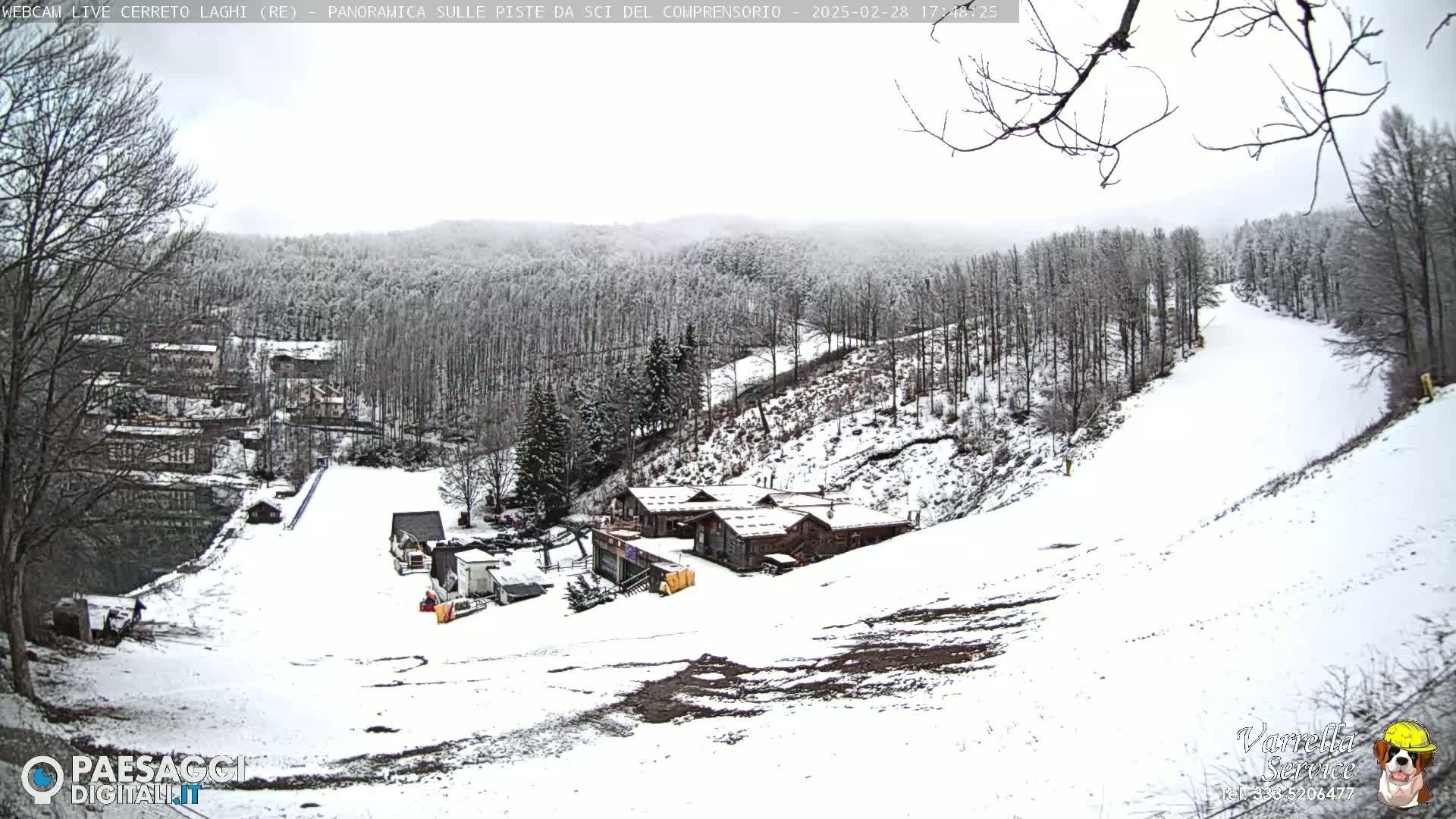 A snow-covered ski slope with several buildings and a wooded hillside in an overcast sky.
