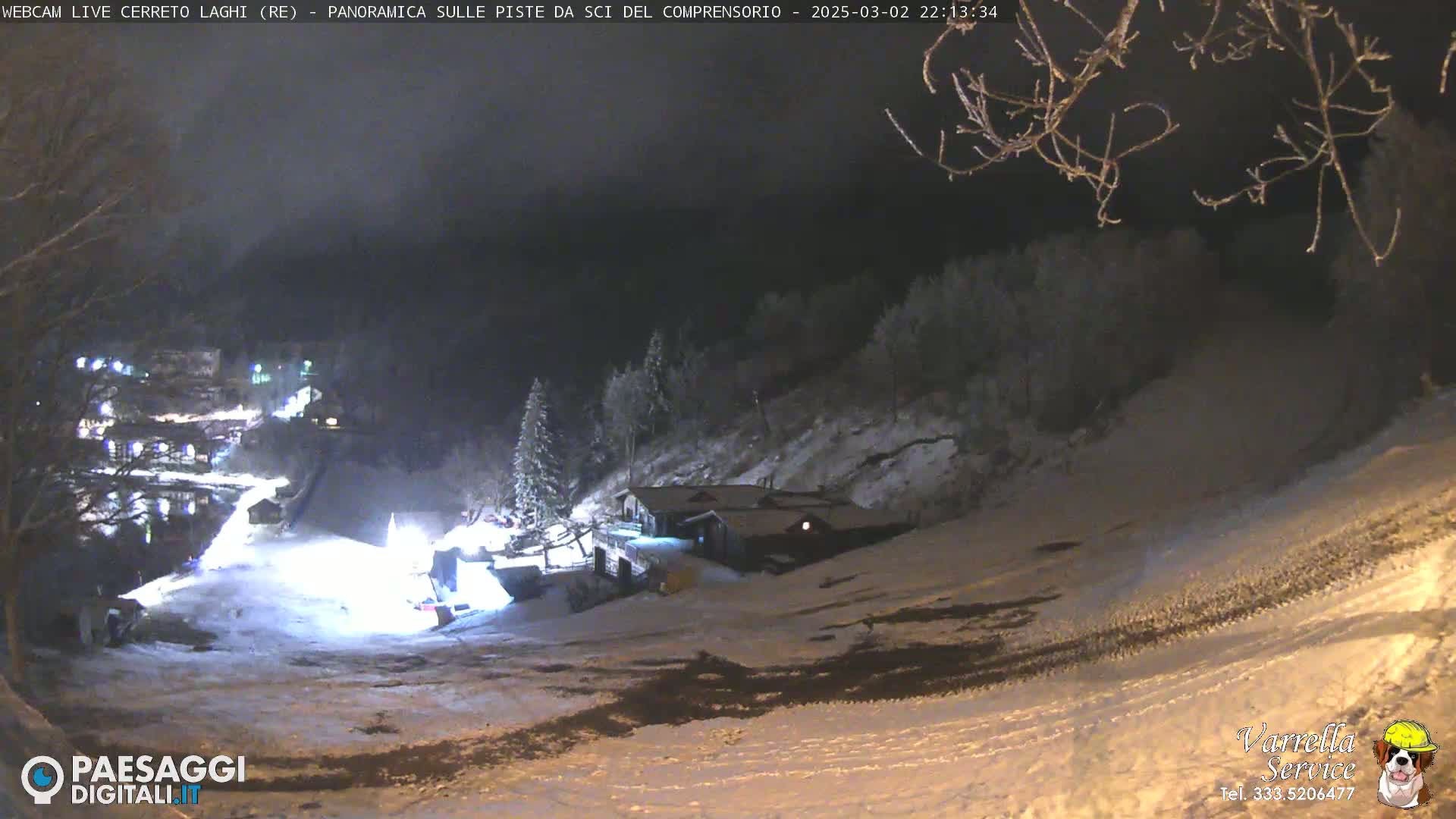 A snow-covered ski slope at night, illuminated by artificial lights, with a building visible on the hillside and trees in the background.