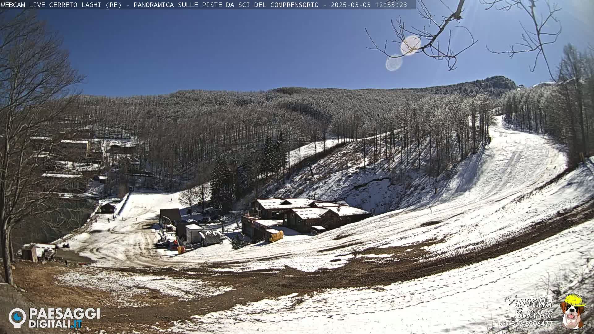A sunny day at a snow-covered ski resort, showing a partially snow-covered ski slope, several buildings, and a largely wooded hillside.