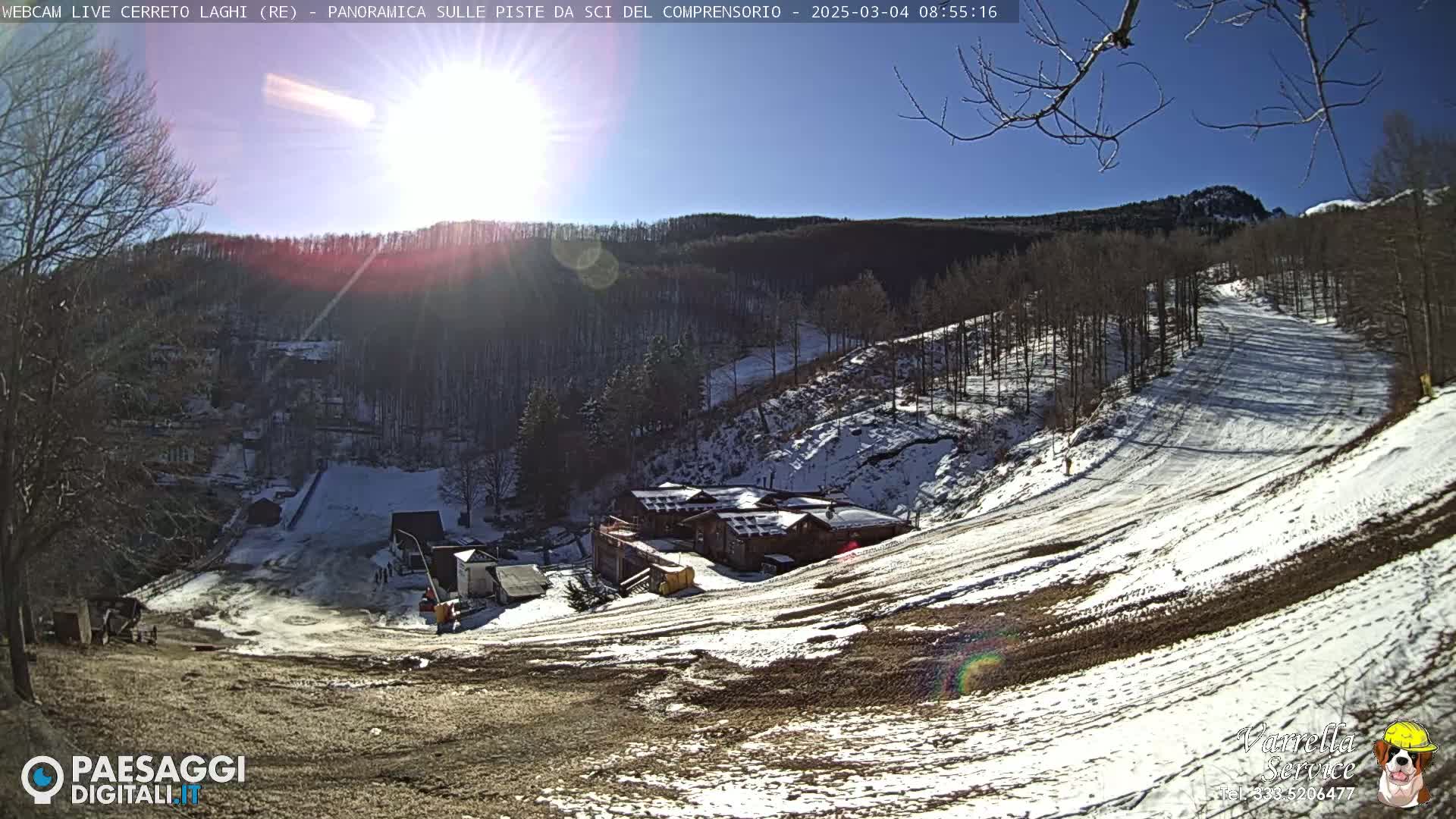 A sunny day at a snow-covered ski resort shows a building complex nestled among sparsely snow-covered trees and ski slopes.