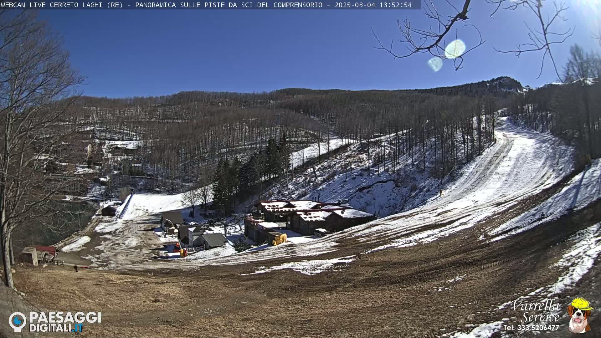A sunny day reveals a snow-covered ski slope with a few buildings at its base and a wooded hillside in the background.