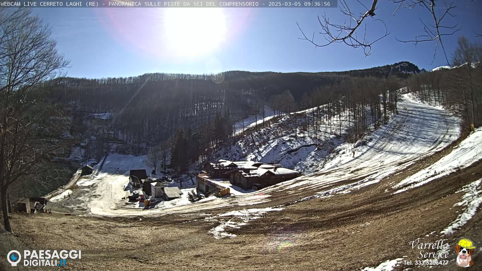 A sunny, snow-dusted ski slope descends from a building complex nestled among sparsely snow-covered trees and hills.