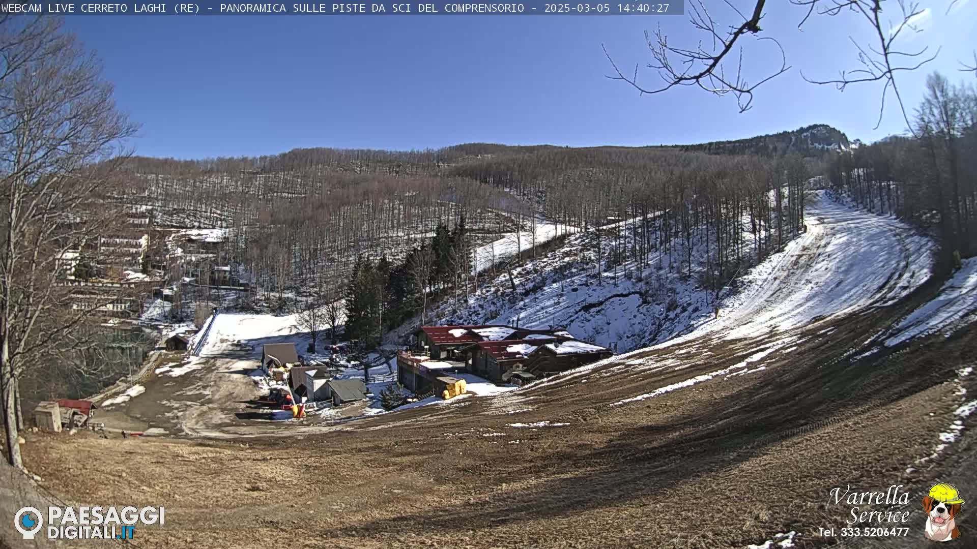 A sunny, partially snow-covered ski slope is viewed from above, showing a ski lodge and surrounding buildings nestled among bare trees.
