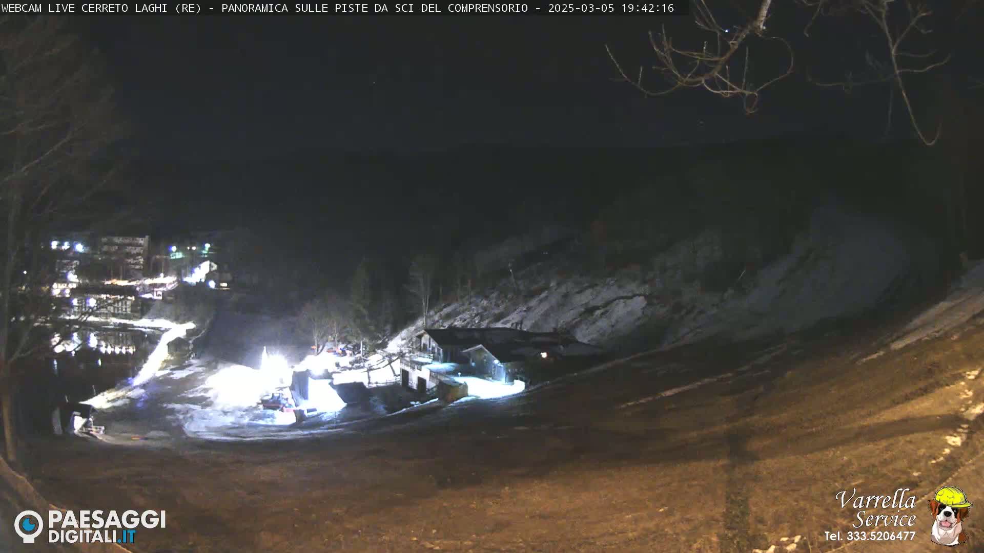 A nighttime, snow-covered ski slope is illuminated by artificial lights, with buildings and trees visible in the distance.