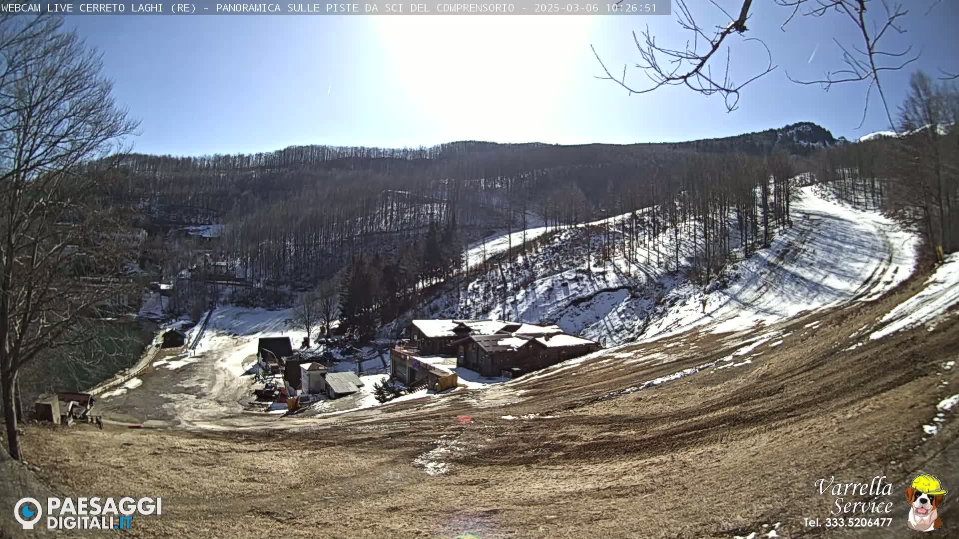 A sunny, snow-dusted ski slope descends toward a cluster of buildings nestled in a wooded valley.