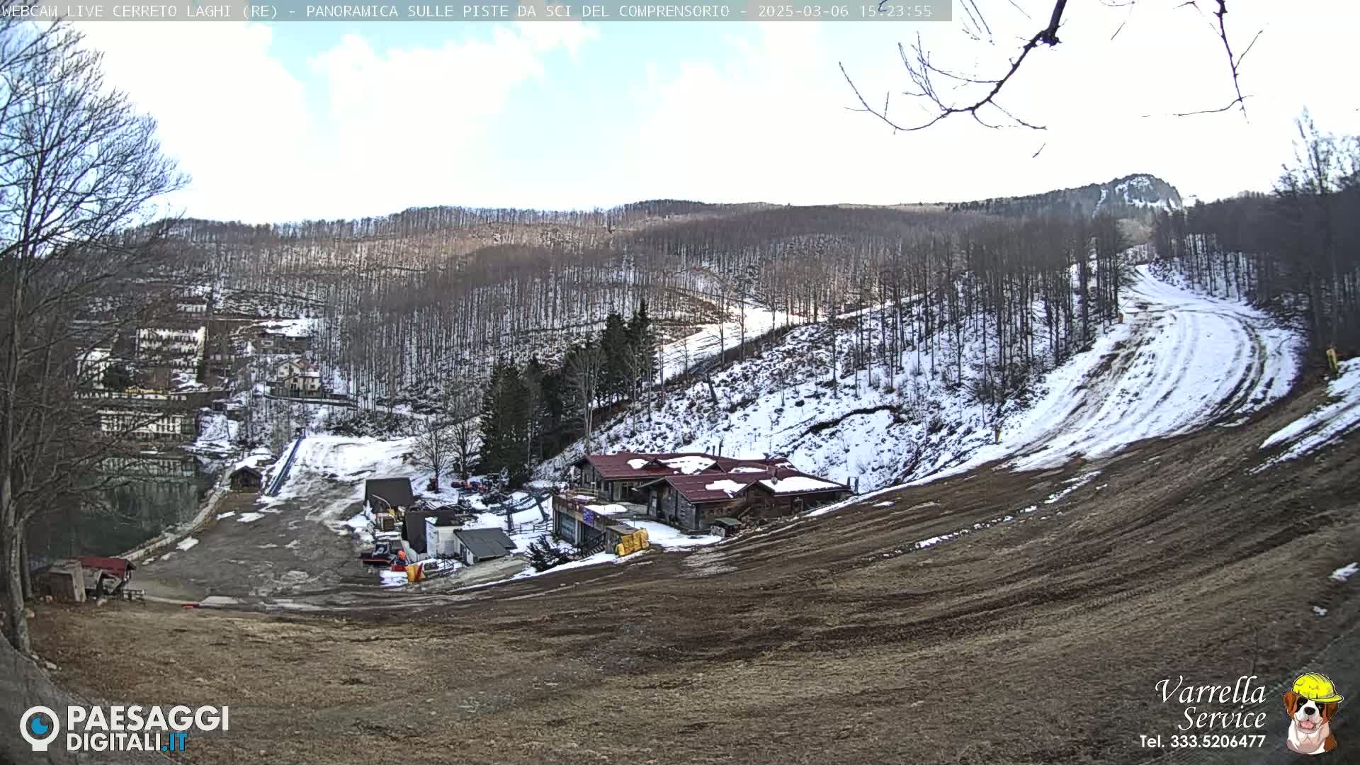 A mostly clear day at a ski resort shows a partially snow-covered hillside with several buildings and ski runs, some of which are bare earth.