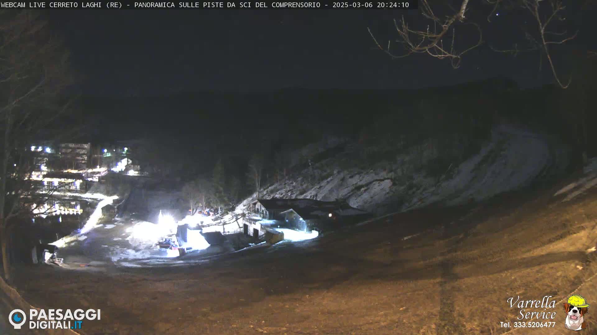 A nighttime, snow-covered ski slope is illuminated by artificial lights, with a building visible in the midground.