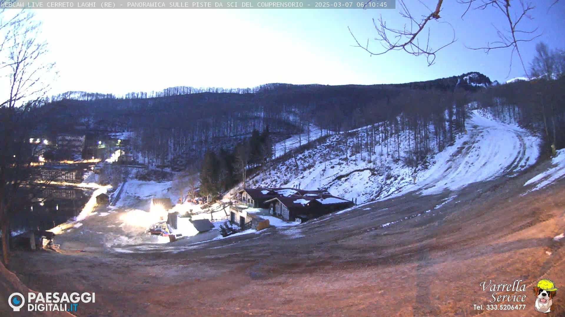 A twilight view of a snow-covered ski slope with buildings and sparsely populated wooded hills in the background.