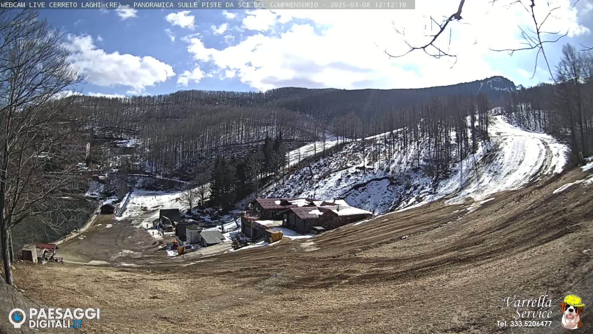 A mostly sunny day overlooking a snow-dusted ski slope with buildings at the bottom of the hill and bare trees on the mountainside.