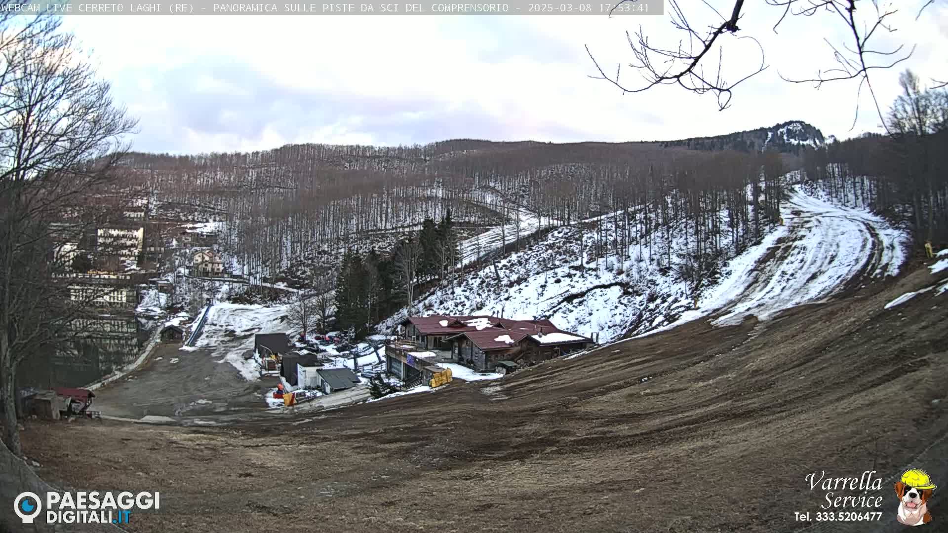 A mostly cloudy day reveals a snow-dusted ski slope with a building complex and sparse trees in the background.
