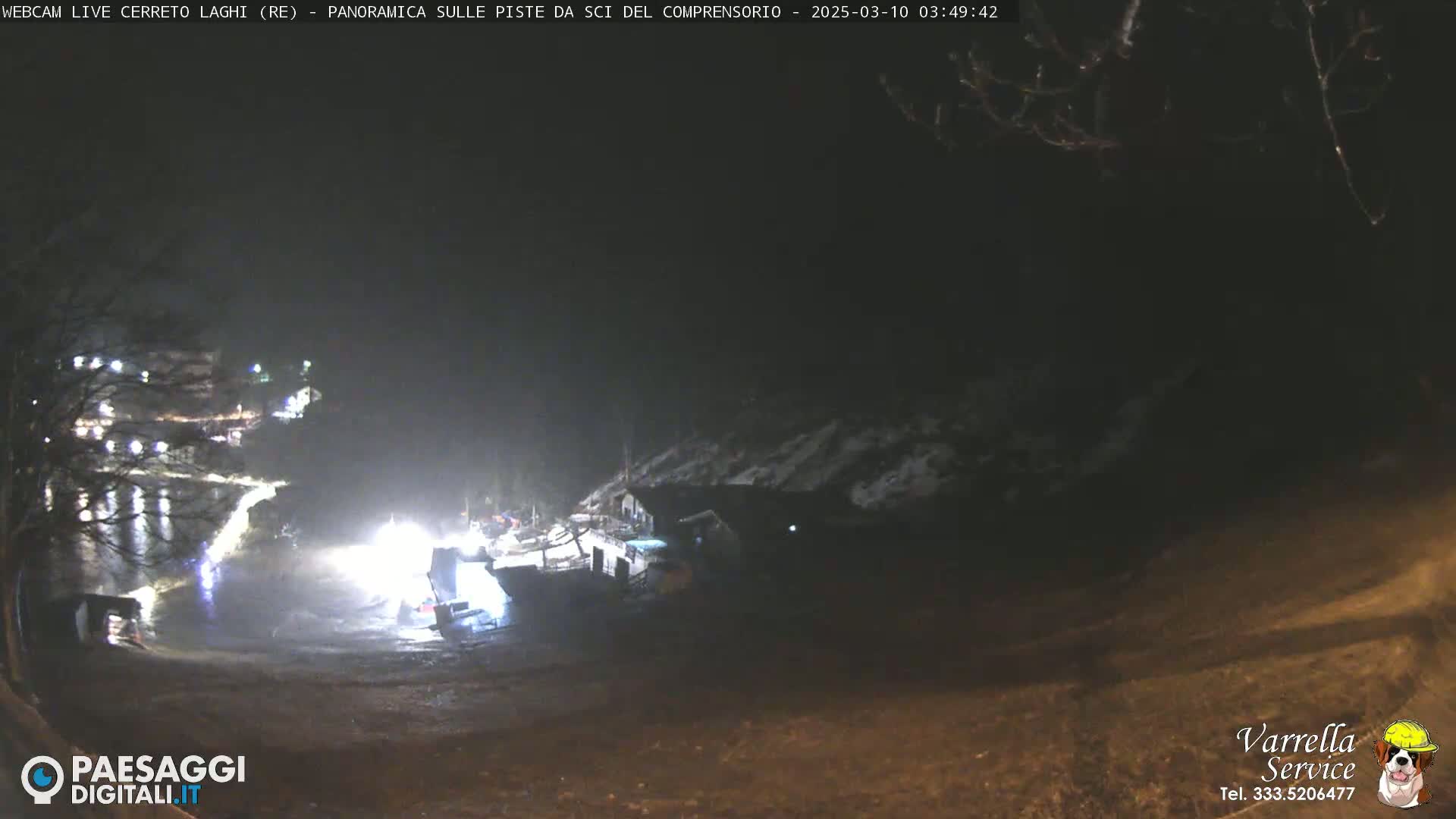 A nighttime, low-light view of a snow-covered landscape with illuminated buildings and vehicles near a body of water.
