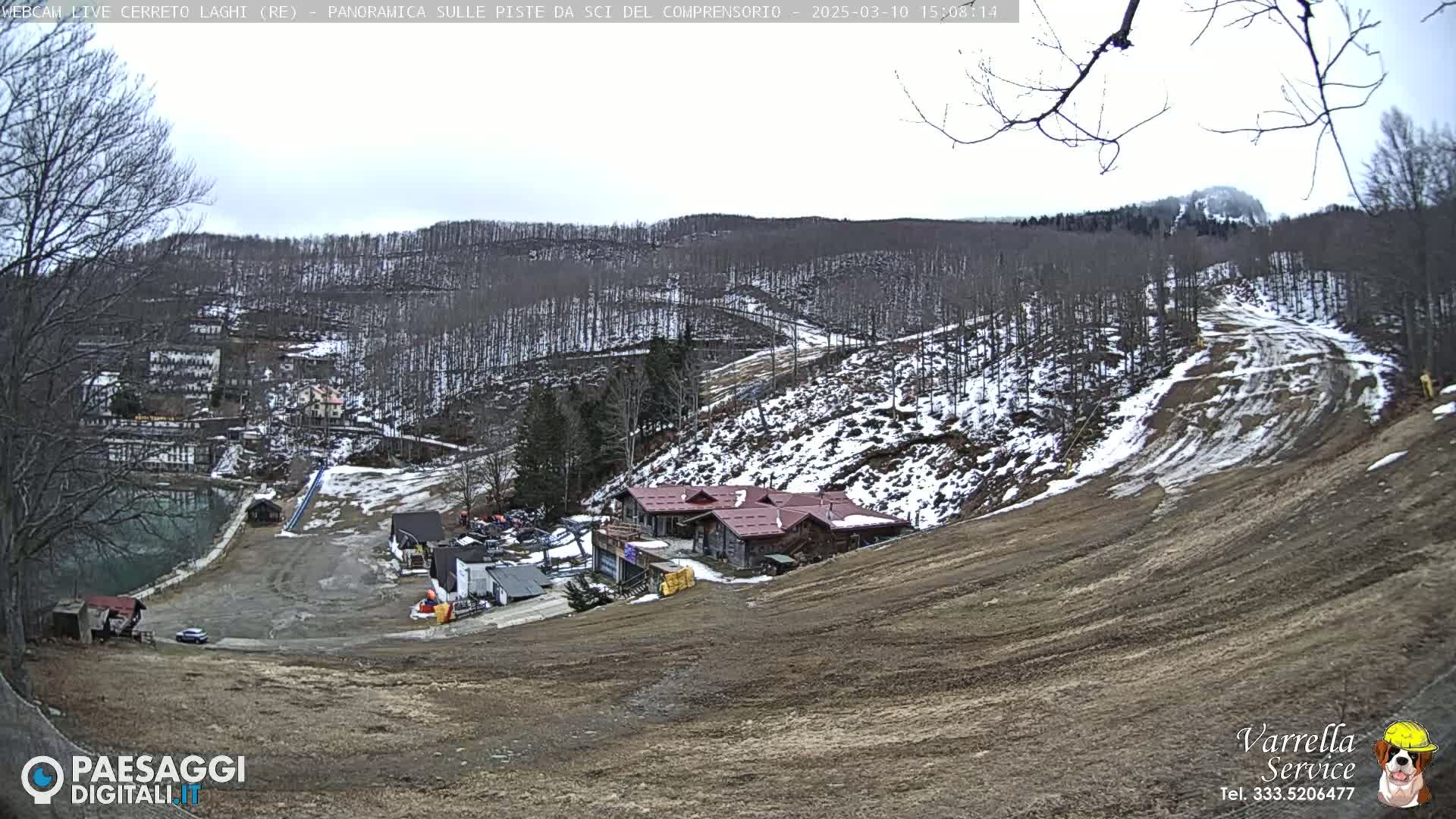 A mostly cloudy day overlooks a ski resort with a brown, snow-dusted slope, a few buildings, and a small lake.