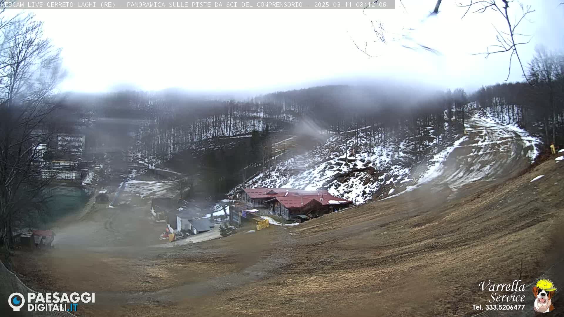 A hazy, overcast view of a snow-dusted ski slope with a building complex and a small body of water in the distance.
