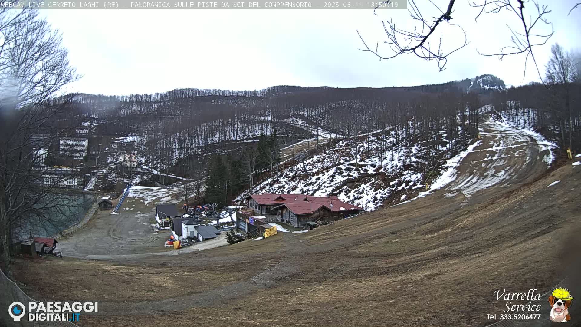 A mostly clear day overlooking a ski resort with some snow on the slopes and surrounding hills, a lodge situated near the bottom of a hill, and bare trees.