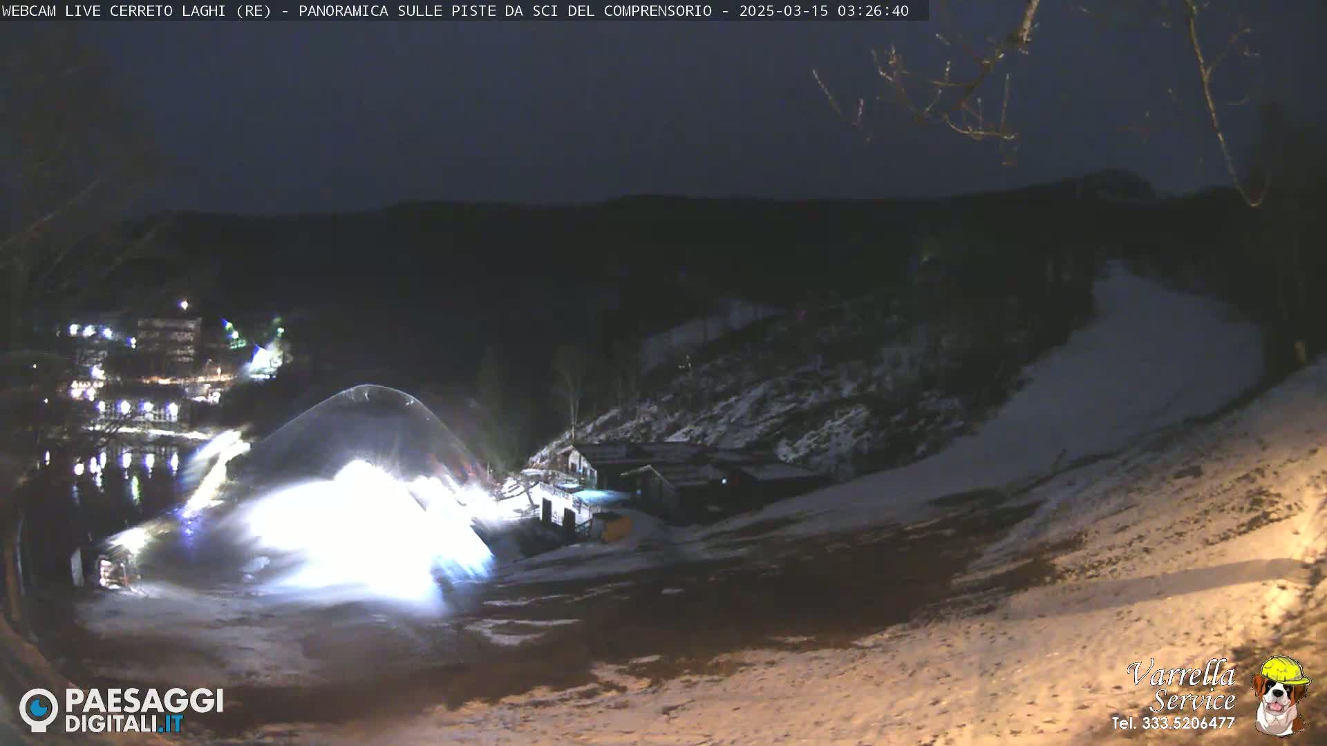 A snowmaking machine illuminates a snow-covered ski slope at night.