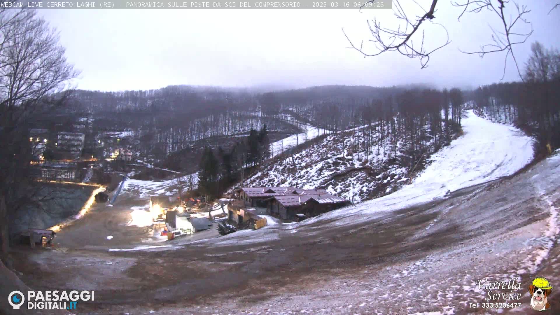 A mostly cloudy, overcast view of a snow-dusted ski slope with a few buildings at the bottom of the hill, and a small town visible in the distance.
