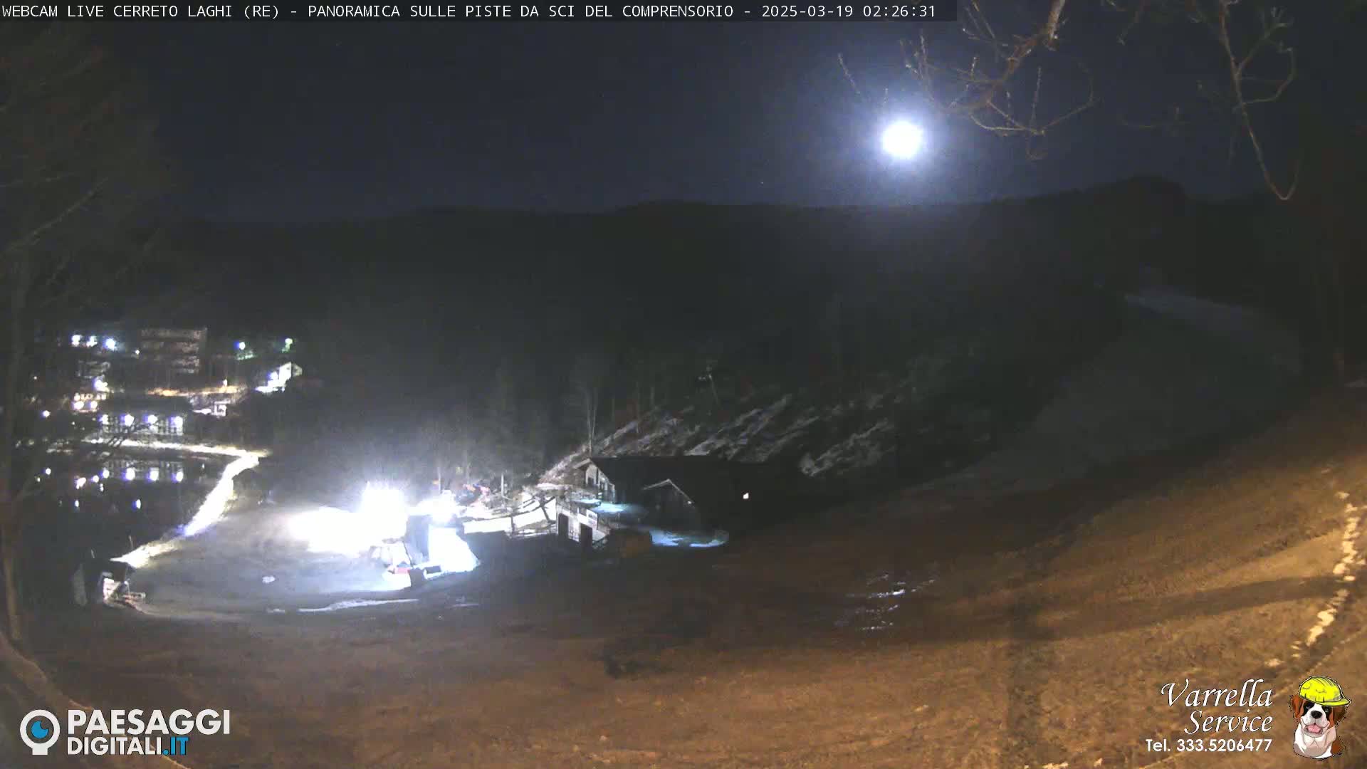 A nighttime, moonlit view of a snow-covered ski resort with a building illuminated near a dark, sparsely treed hillside.