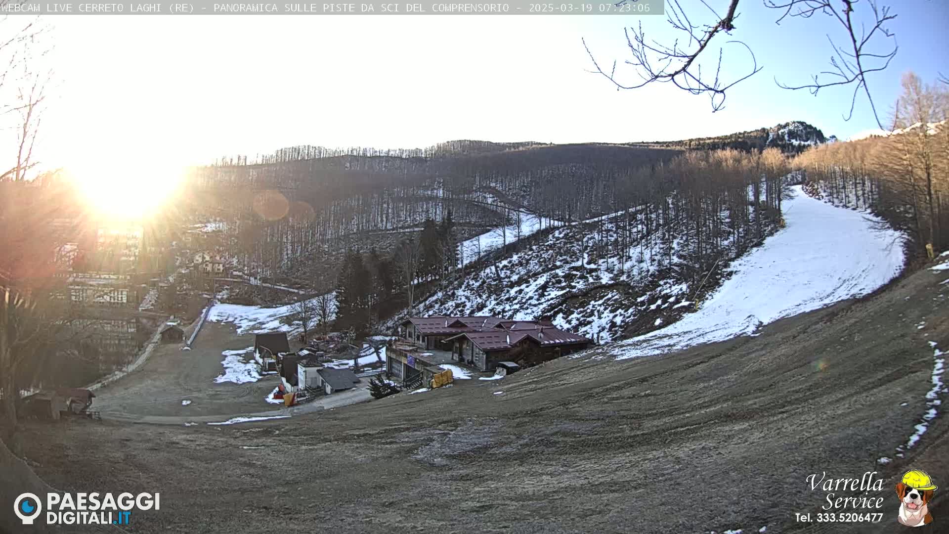 A sunny, wintery scene shows a ski slope with a few buildings at the bottom of the hill, partially covered in snow and surrounded by mostly bare trees.