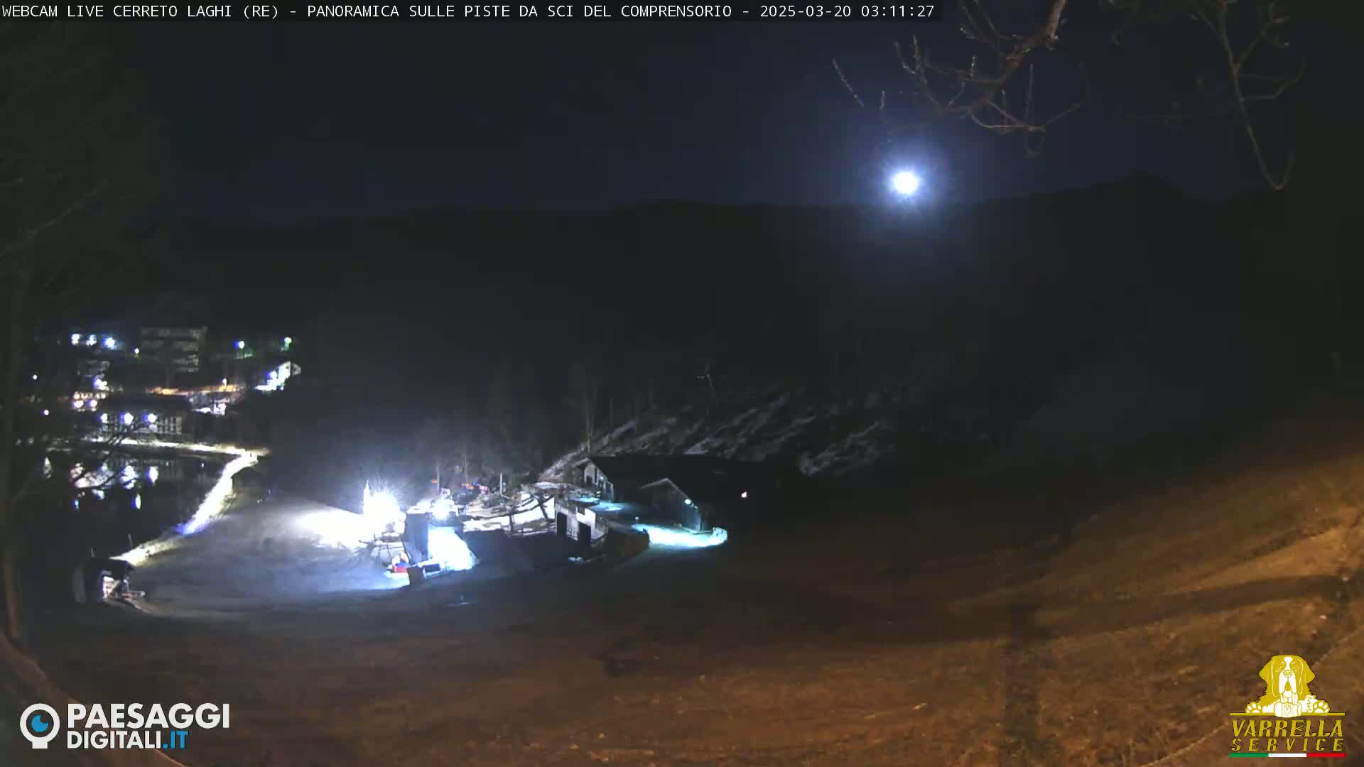 A nighttime view of a snow-covered ski resort illuminated by artificial lights under a bright moon.