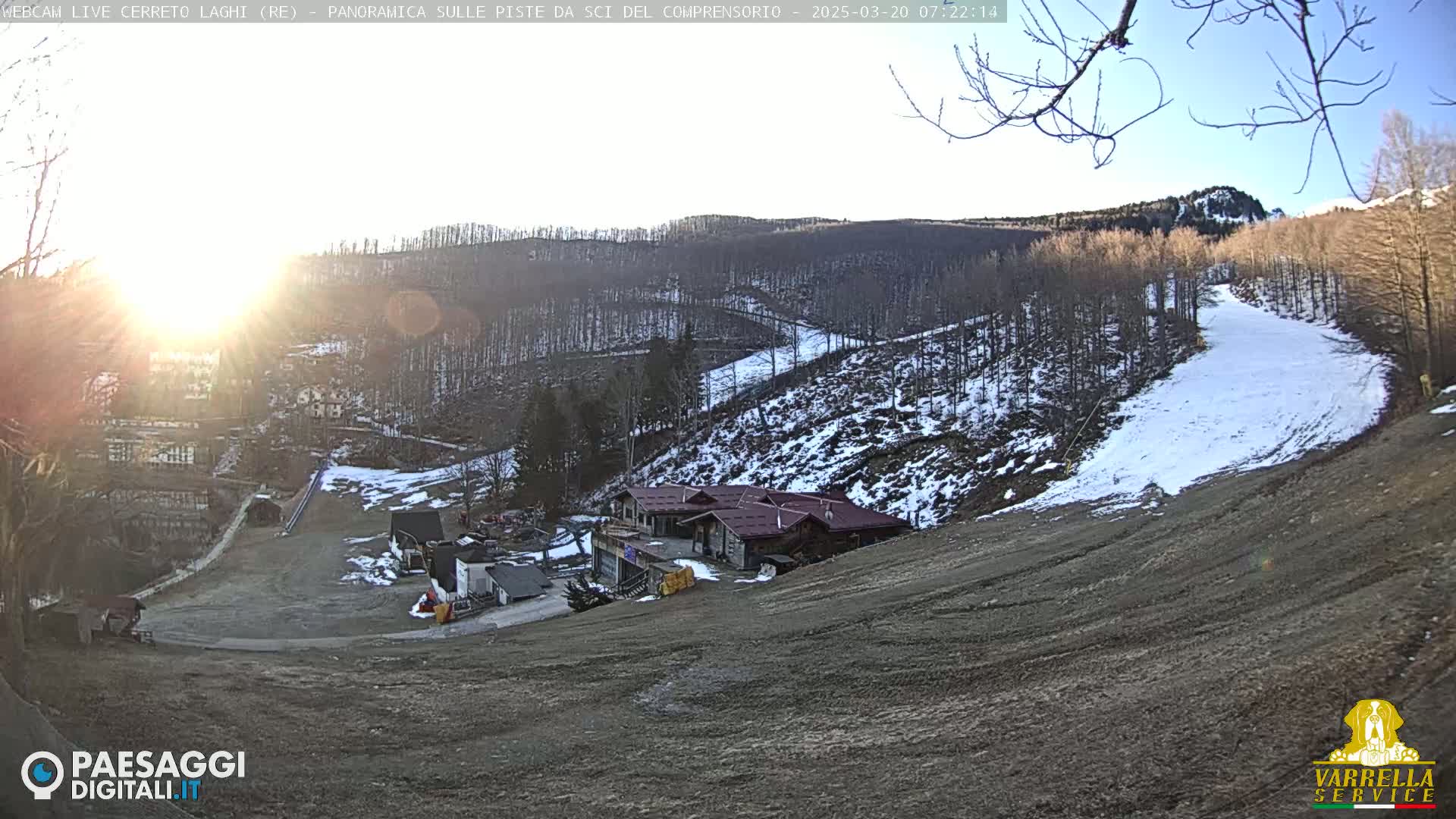 A sunny, wintery scene shows a ski slope with a building at its base, surrounded by bare trees and patches of snow on a hillside.