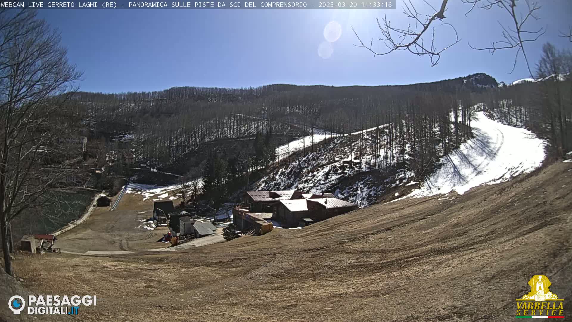 A sunny, snow-dusted ski slope descends towards a small cluster of buildings nestled in a wooded mountain valley.