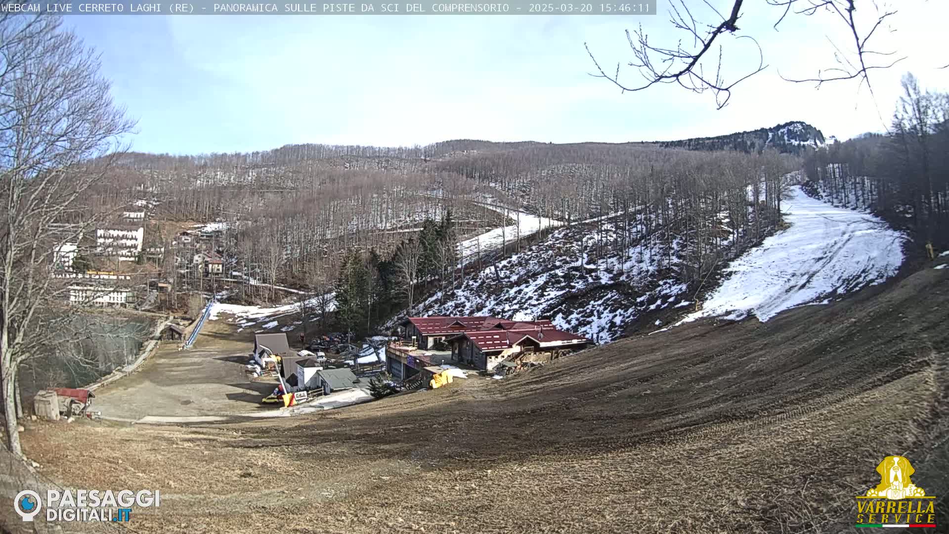 A sunny view of a ski resort with partially snow-covered slopes and several buildings nestled amongst leafless trees.