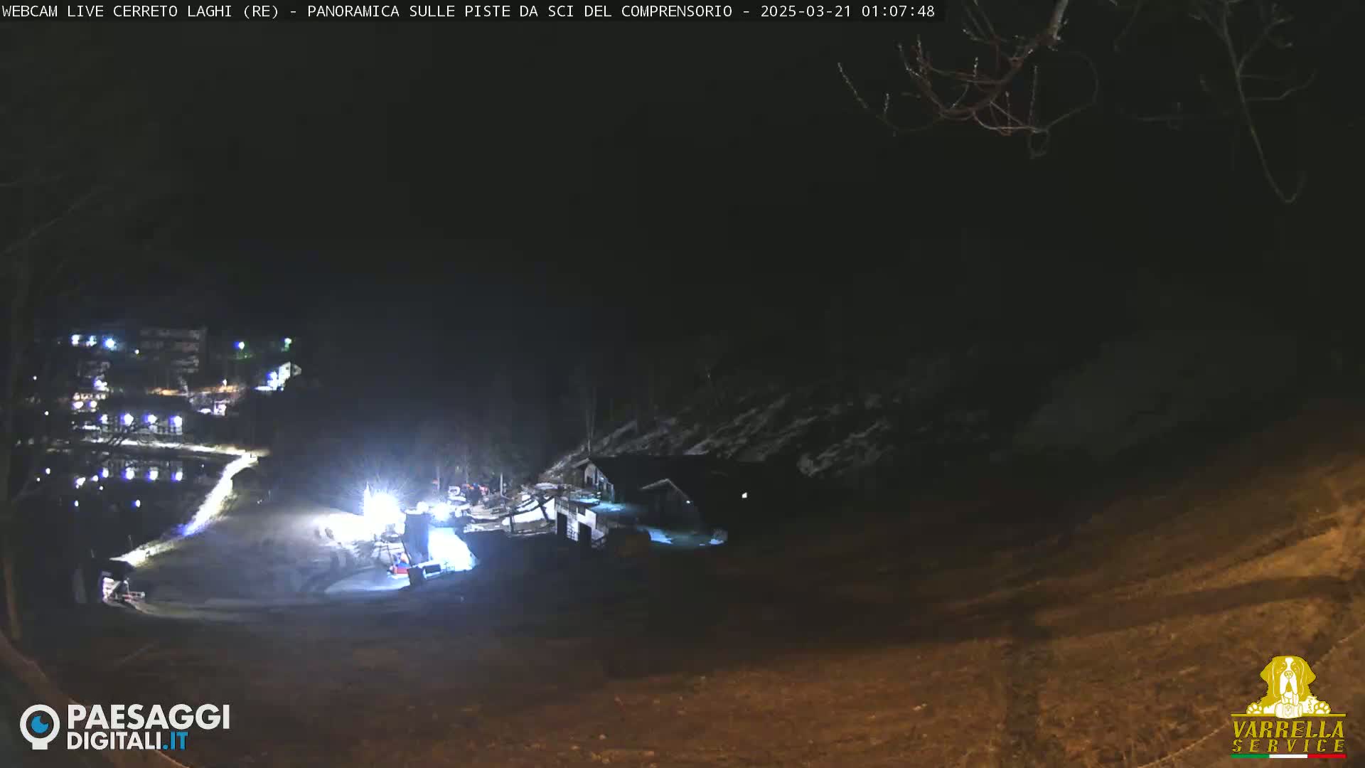 A nighttime, long shot of a snow-covered hillside with a few buildings illuminated, a road winding through the scene, and some activity near the bottom.