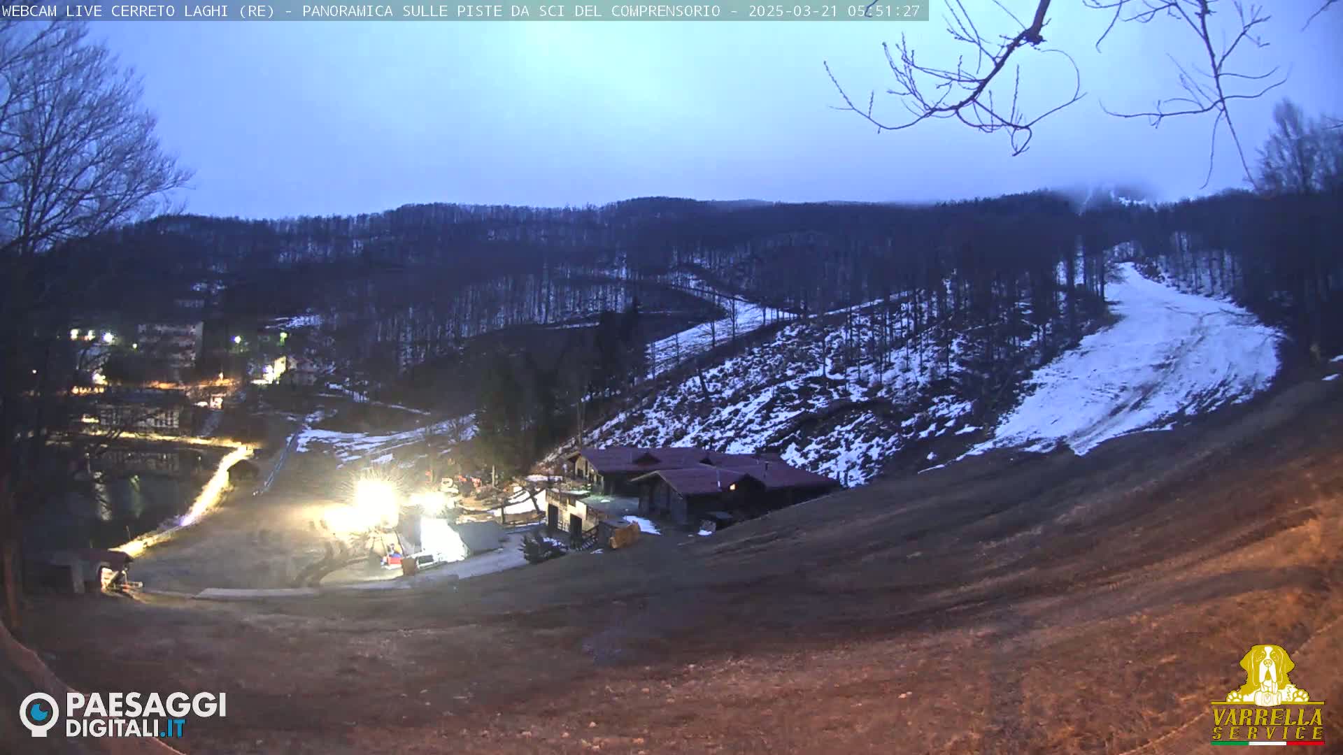 A twilight view of a snow-dusted ski slope and surrounding hills, with lights illuminating a lower area near buildings.