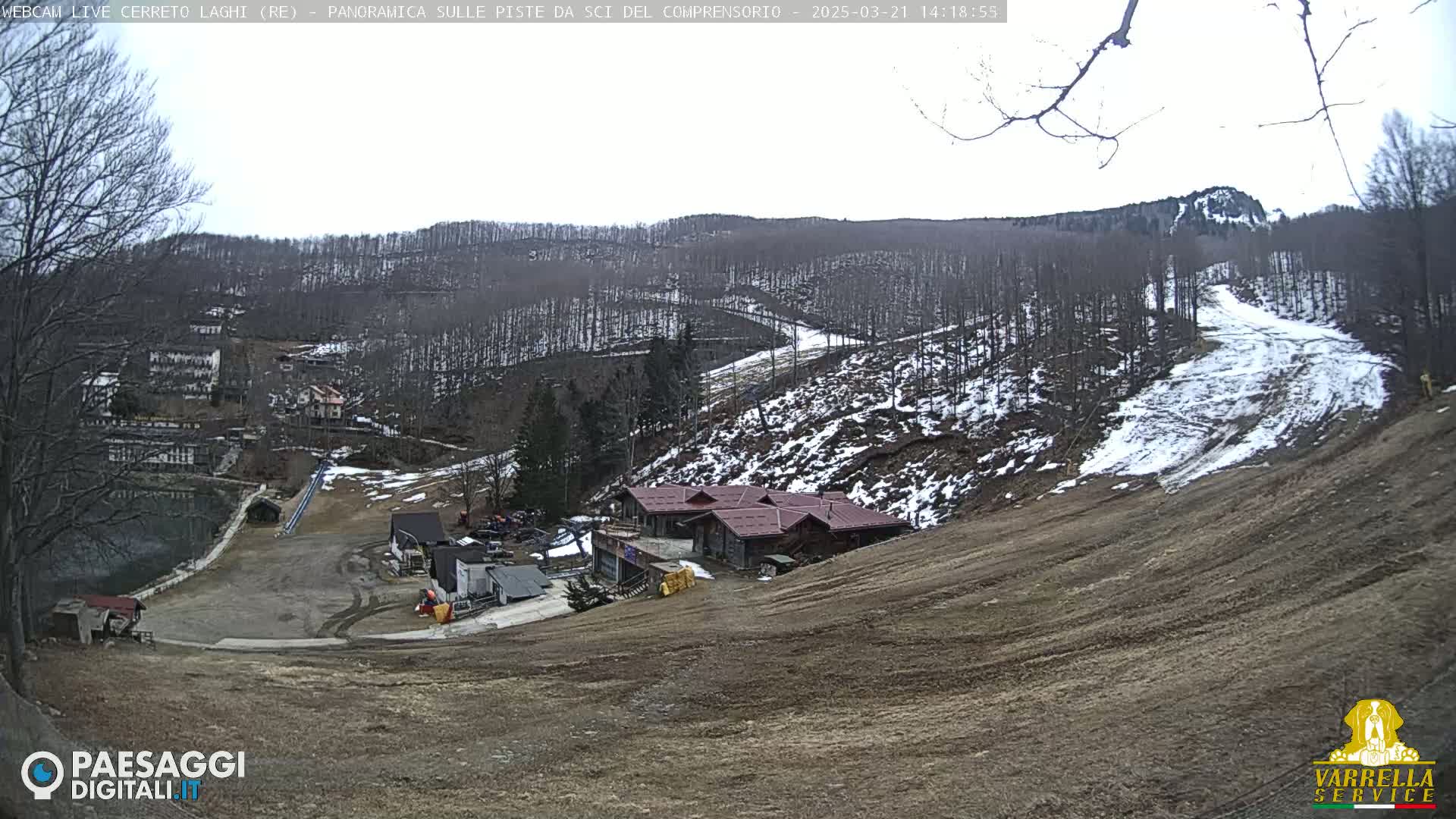 A mostly barren ski slope with patches of snow, a few buildings, and a wooded hillside under an overcast sky.