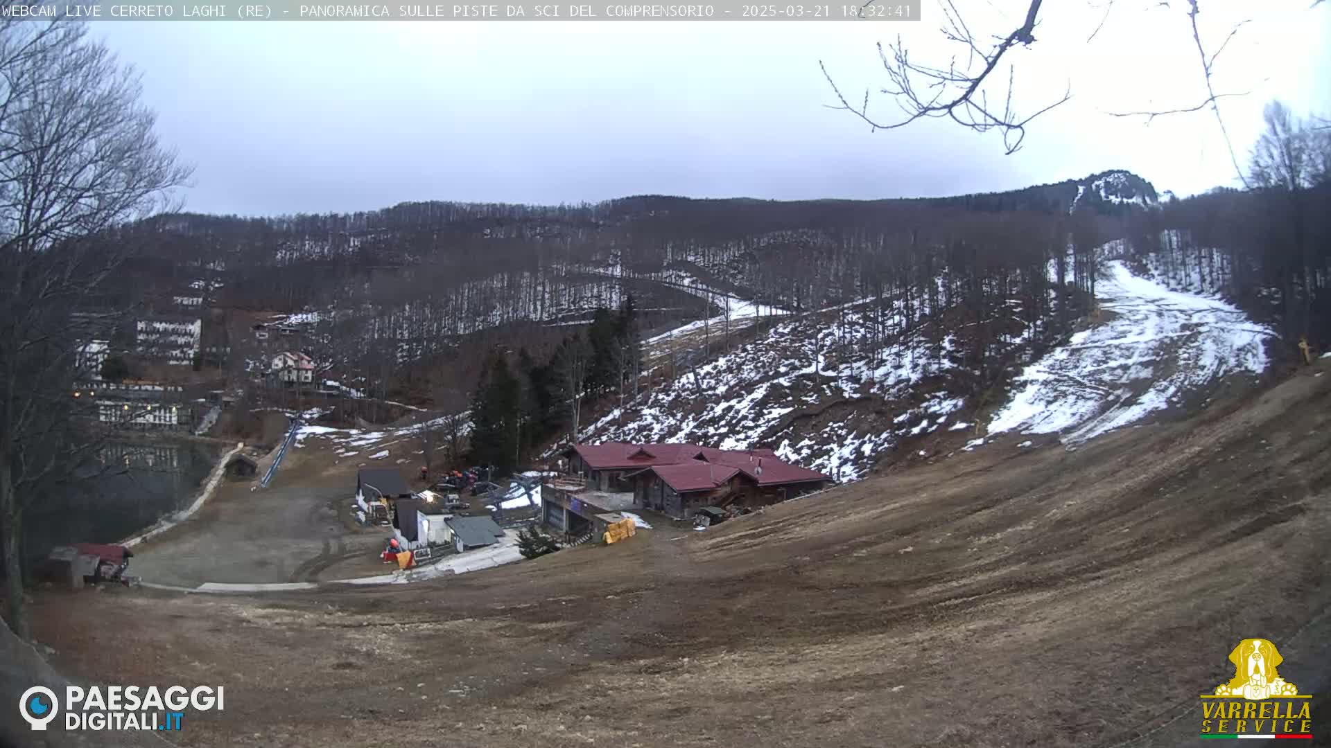 A mostly barren ski slope with patches of snow, a building at its base, and a forested hillside in the background under an overcast sky.