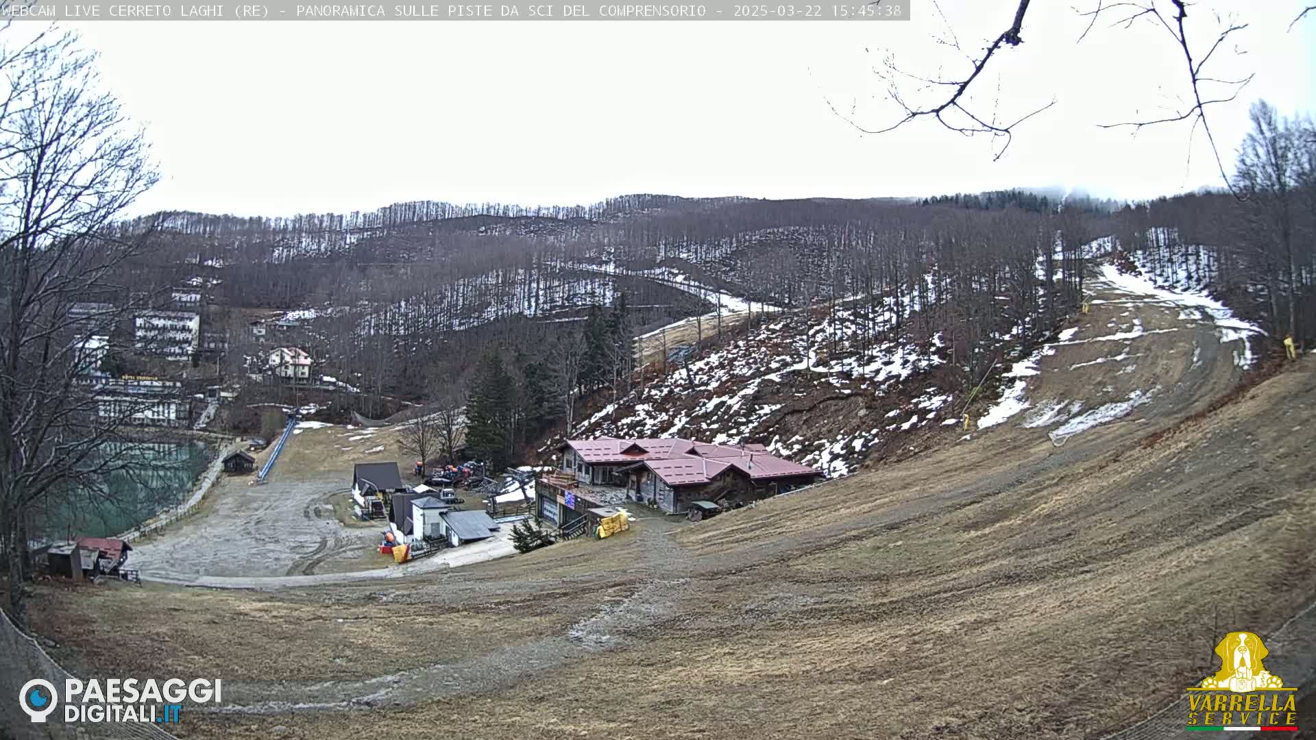 A mostly barren ski slope with a few patches of snow, a building near the bottom, and a forest in the background under an overcast sky.