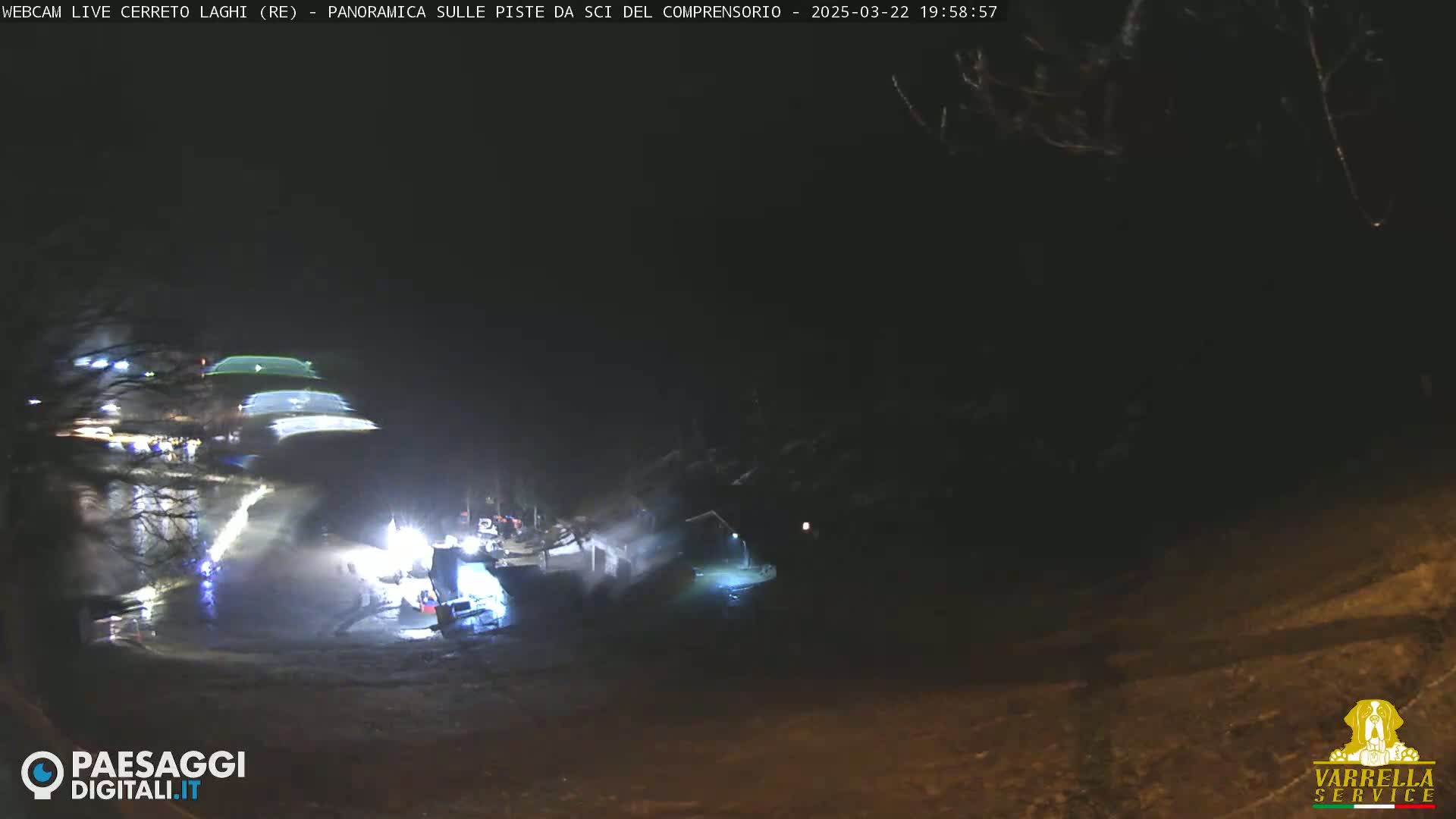At night, brightly lit machinery and vehicles are visible near a dark body of water, surrounded by trees under a dark sky.