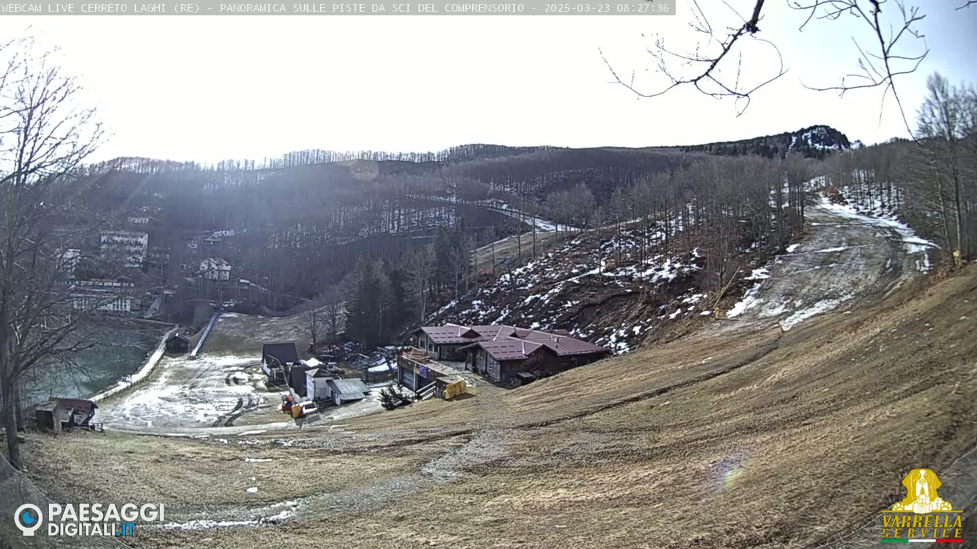 A mountainside ski resort with a lodge, partially snow-covered slopes, and leafless trees under a mostly clear sky.