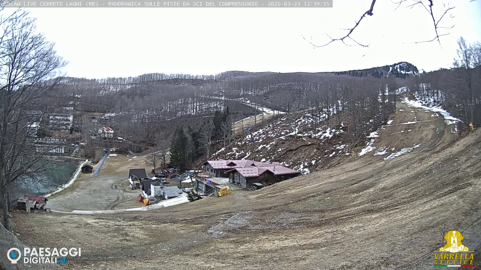 A mostly barren ski slope with a few buildings and a small lake is visible, set against a backdrop of leafless trees and a distant rocky hill under an overcast sky.